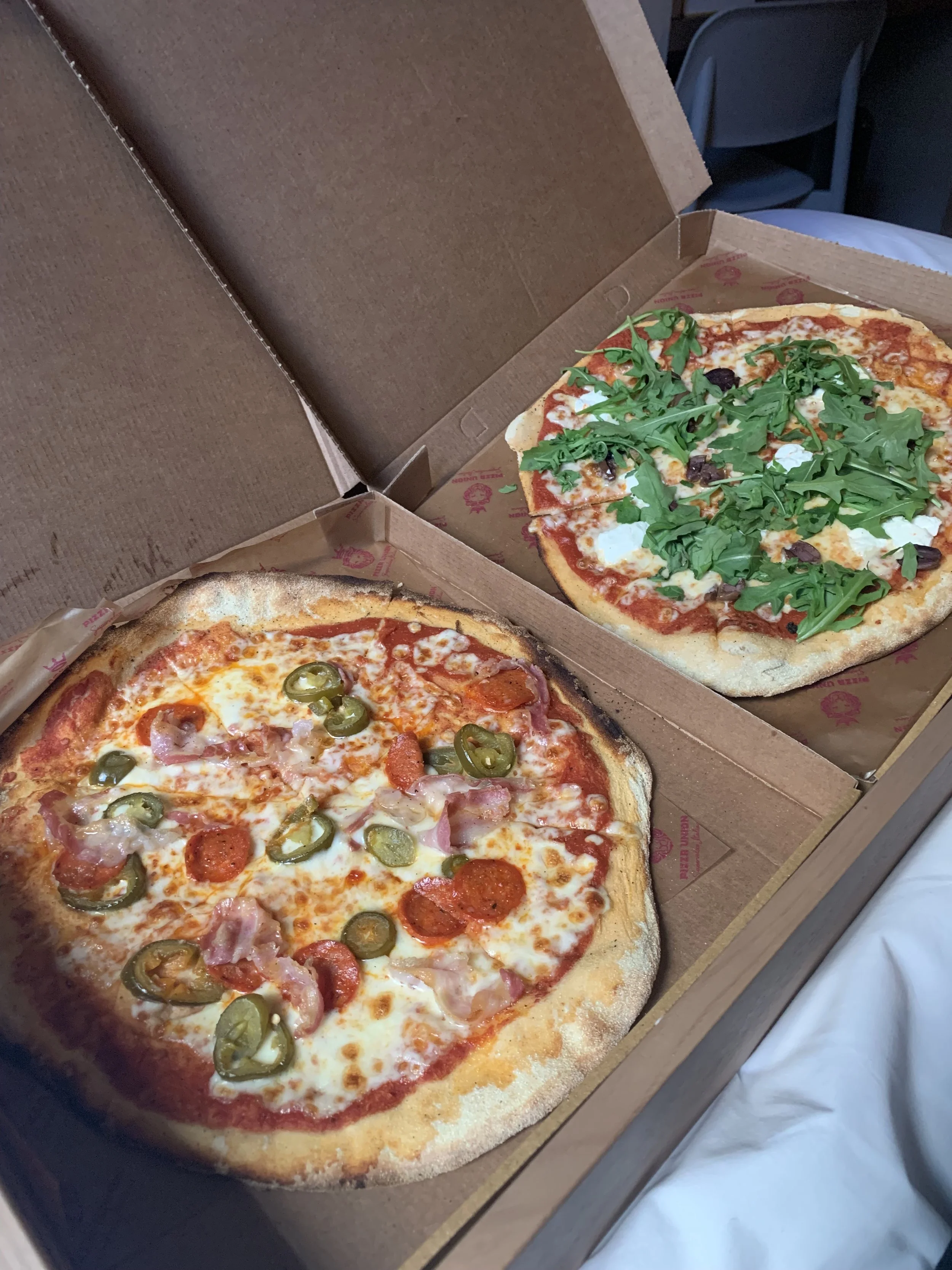 Two pizzas in open brown cardboard boxes, one with pepperoni, green chili slices, ham, and cheese, and the other with cheese, tomato sauce, and arugula.