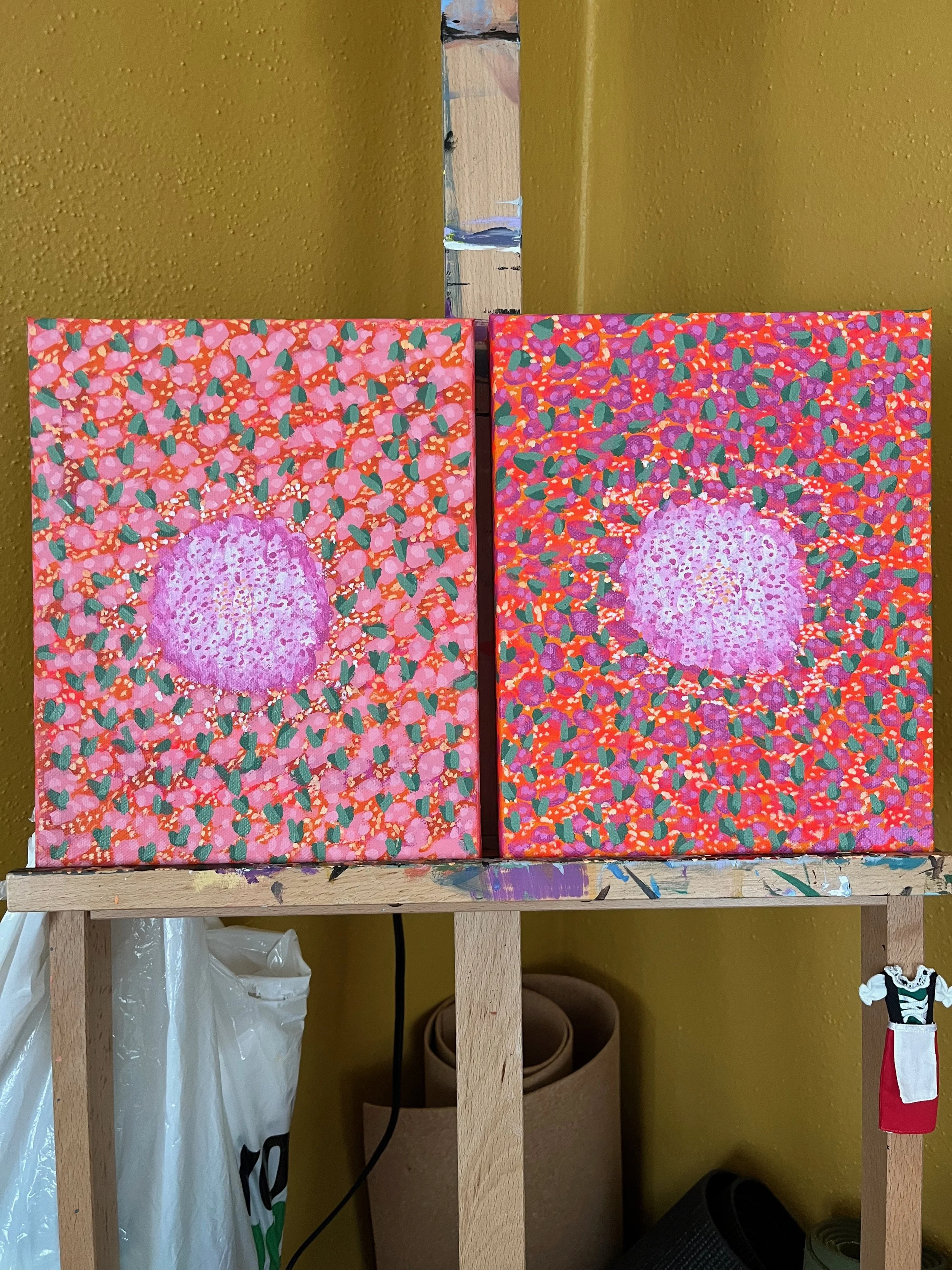 Two colorful abstract paintings with pink, purple, and red circular patterns and green accents, set against a yellow wall on an artist's easel.