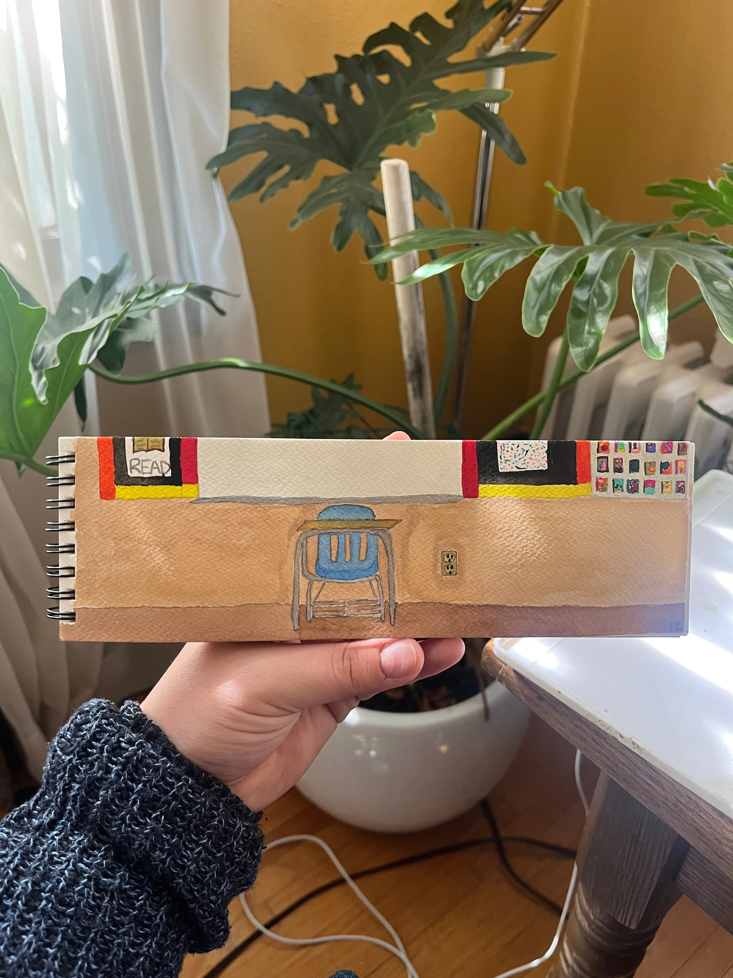 A hand holding a spiral-bound notebook with a painted classroom scene, showing a desk with a blue chair, a whiteboard, and colorful bulletin boards, in front of large green indoor plants near a window.