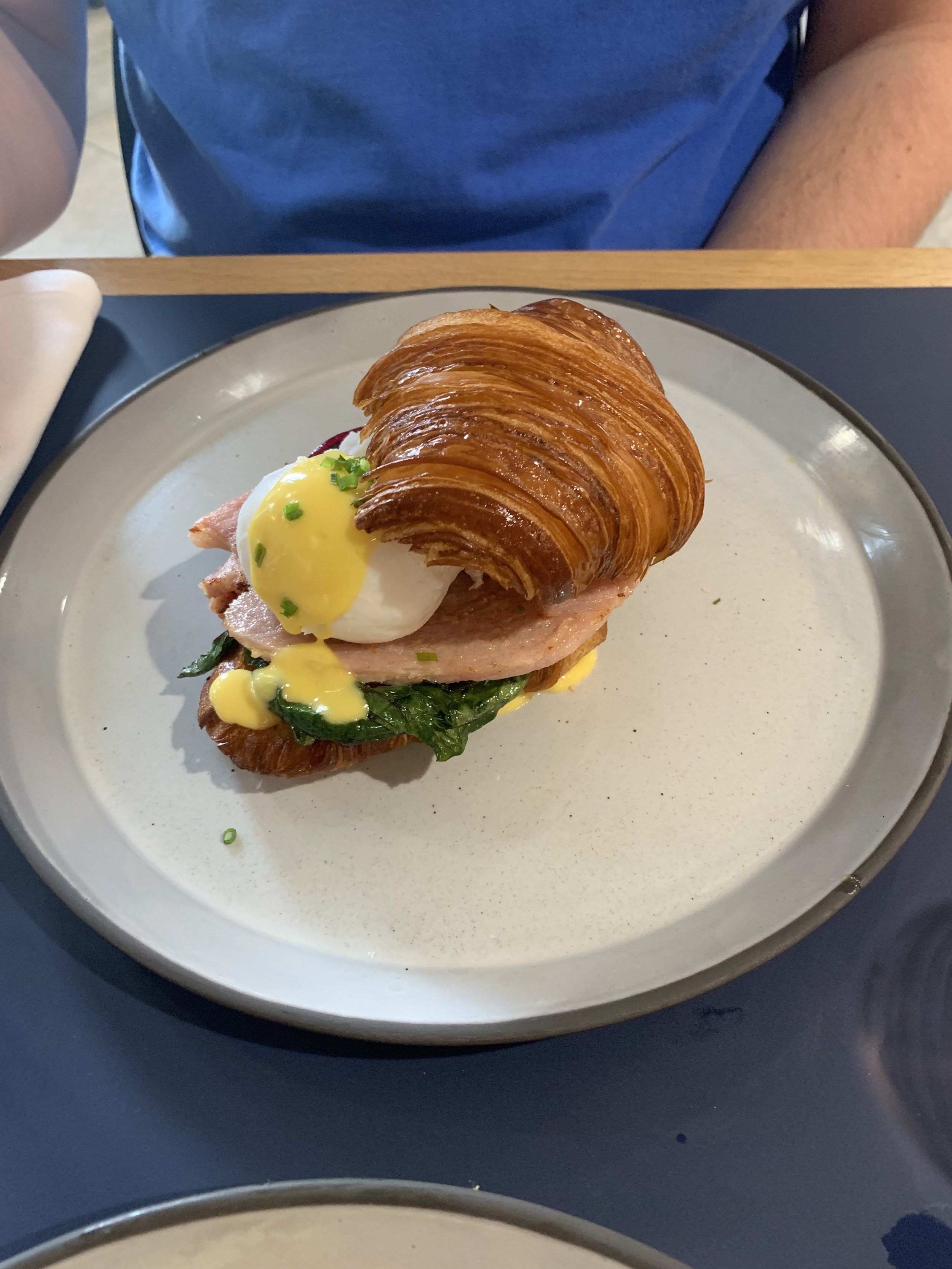 A plate with a croissant sandwich topped with a poached egg, ham, spinach, and yellow sauce.