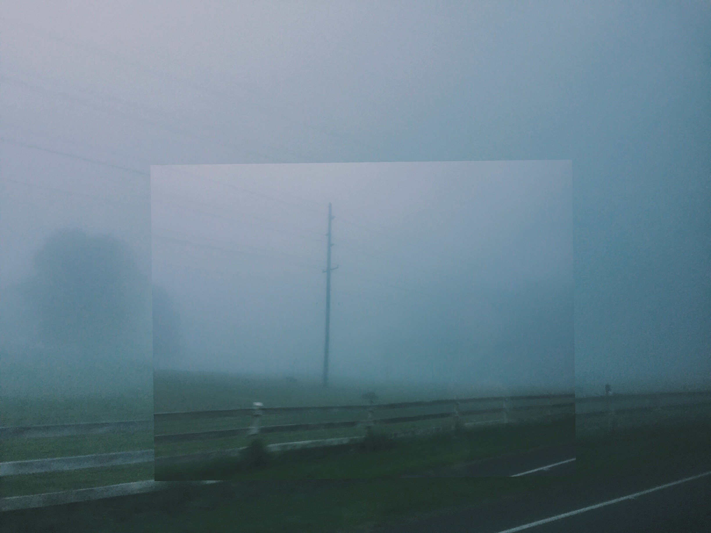 A foggy landscape viewed through a vehicle window with a utility pole, power lines, and a road with a guardrail.