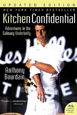 Cover of "Kitchen Confidential: Adventures in the Culinary Underbelly" by Anthony Bourdain, featuring a man in a white chef's coat with crossed arms, standing in front of a dark background with illuminated text.