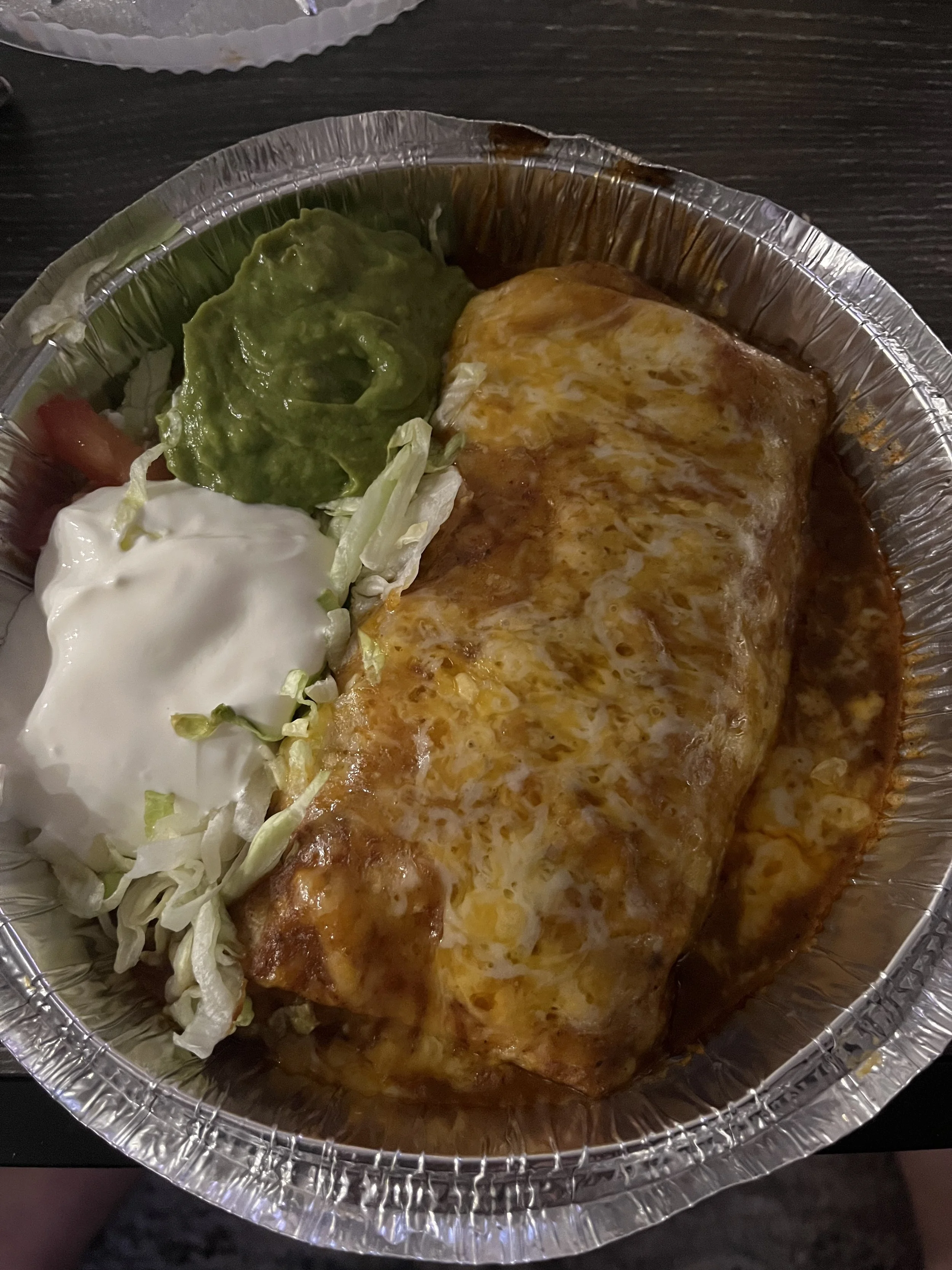 A takeout aluminum tray containing a burrito topped with melted cheese and sauce, with sides of guacamole, sour cream, and shredded lettuce and tomato.