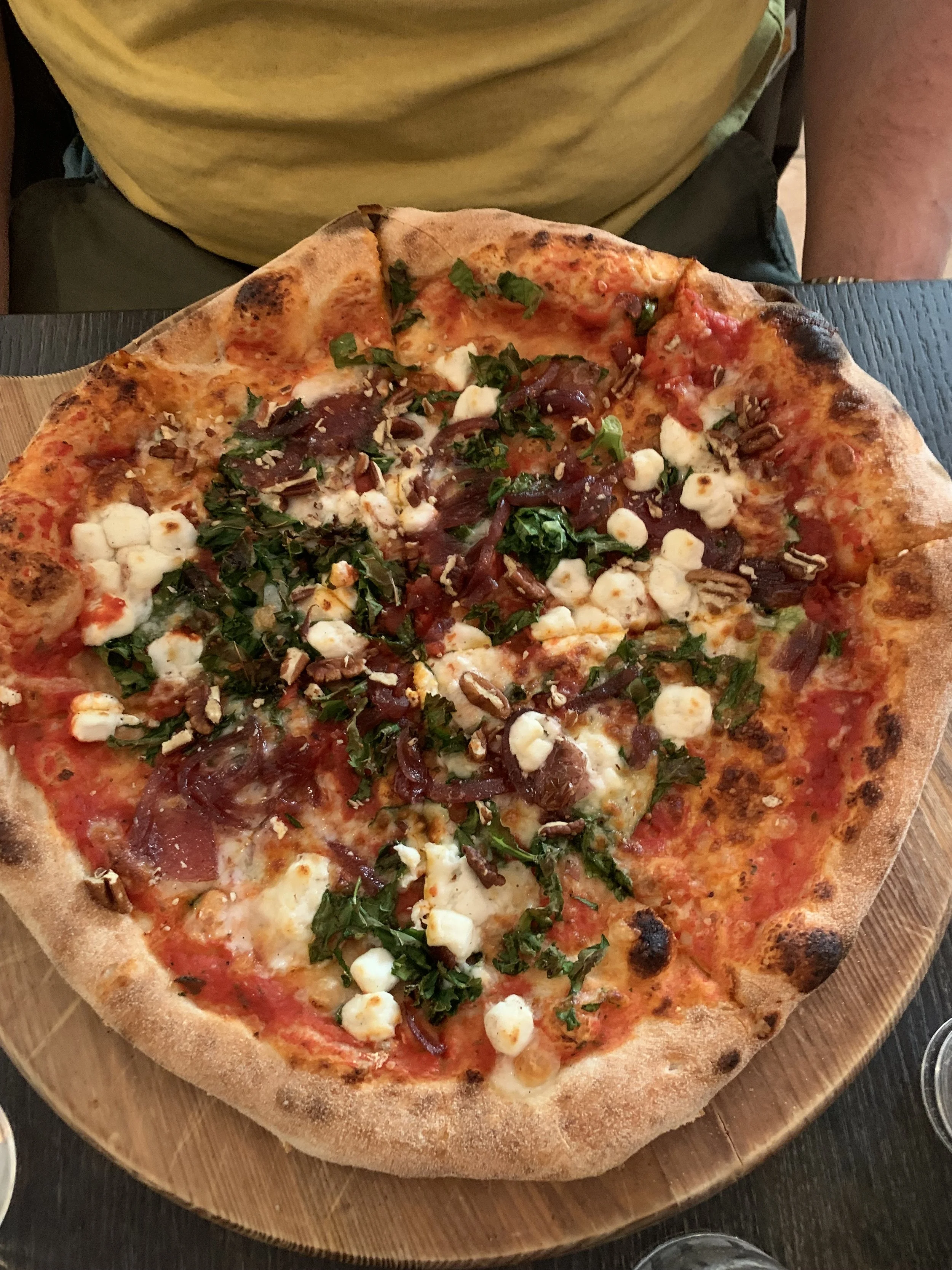 A pizza with a golden crust topped with cheese, spinach, cherry tomatoes, red onions, and crumbled cheese, served on a wooden board.
