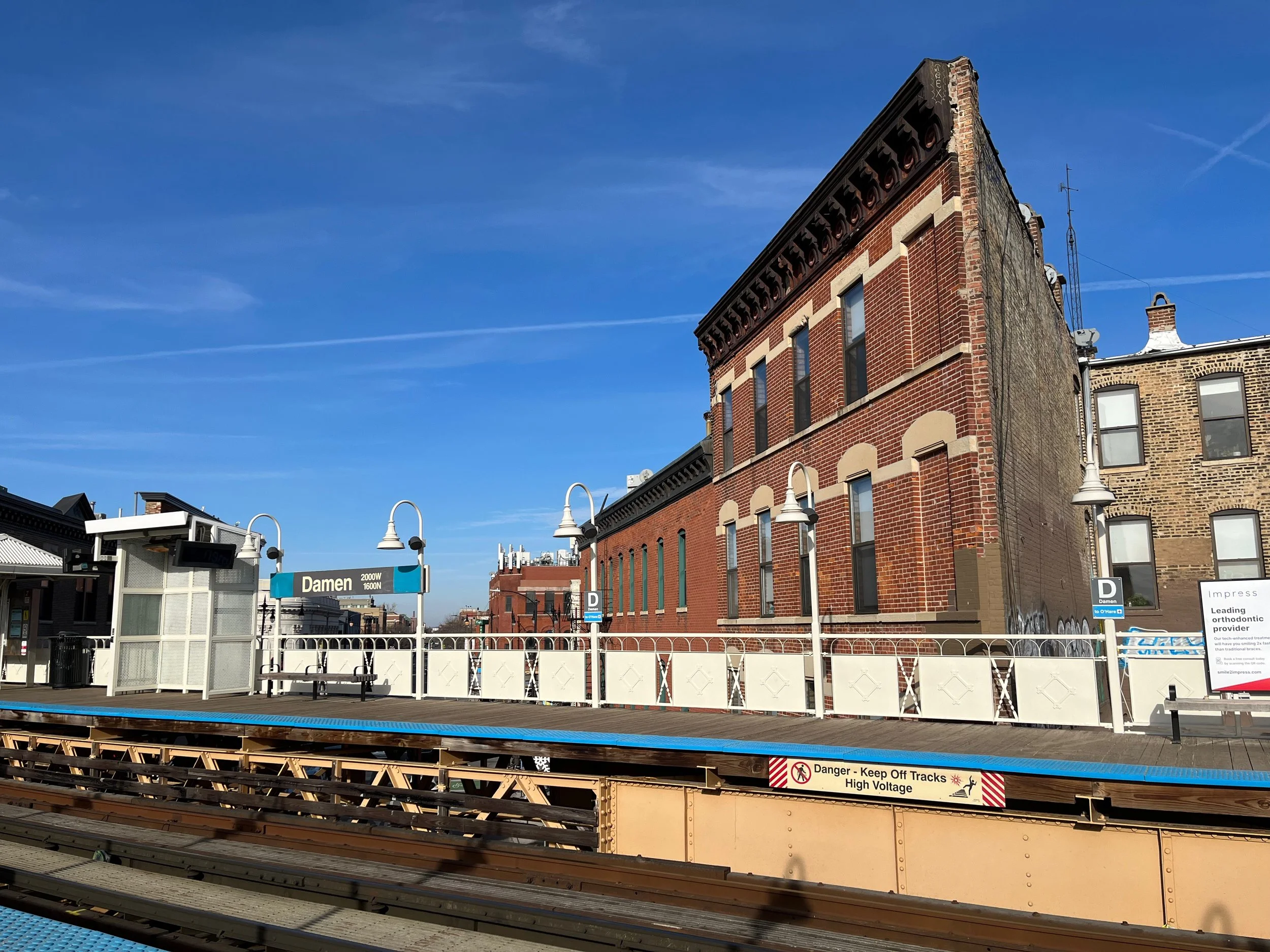 View of Damen train station platform with tracks in the foreground, a sign that reads 'Damen,' vintage brick buildings behind the platform, and a clear blue sky.