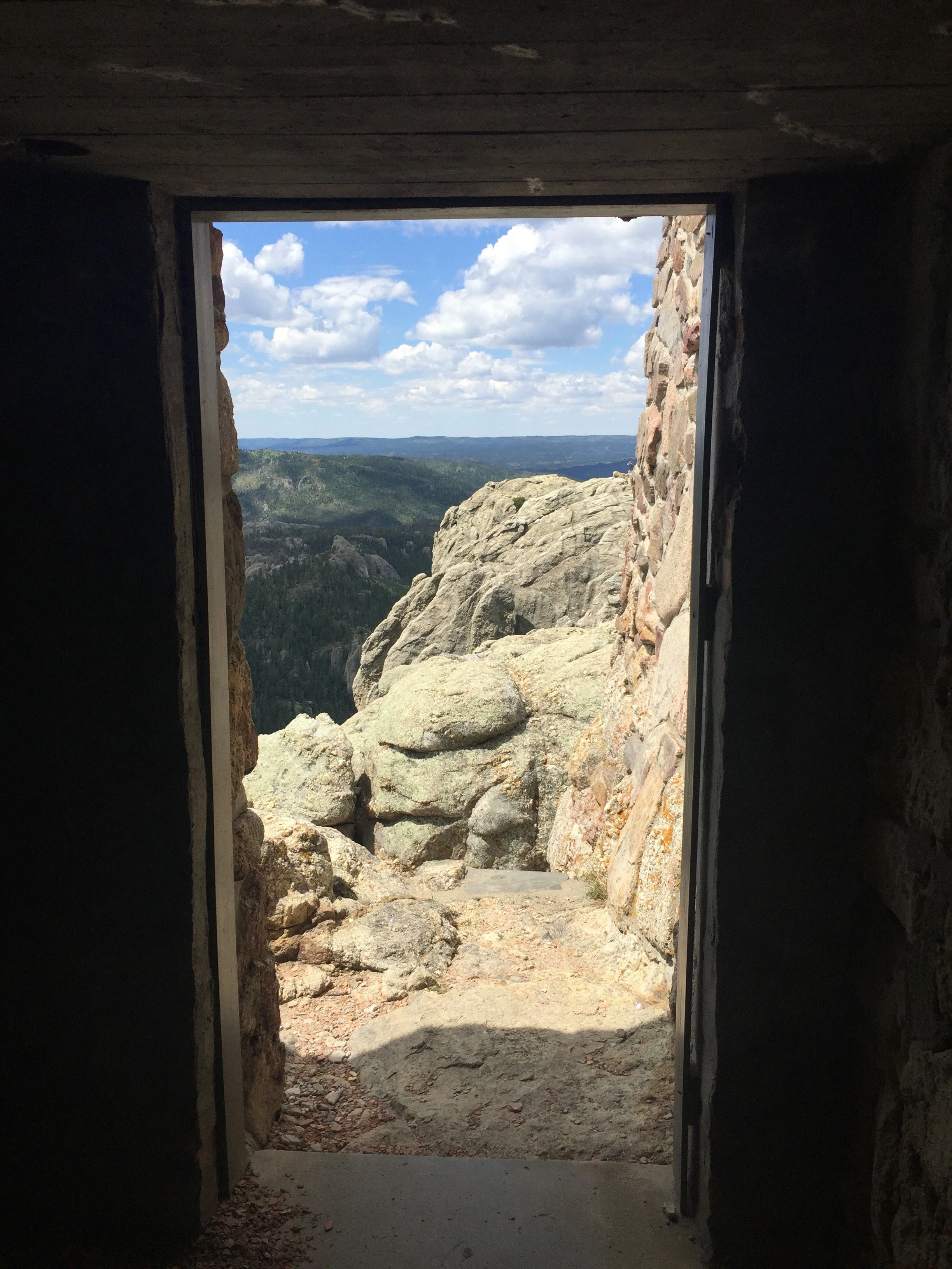 View through a stone-framed opening showing a rocky cliff with a forested valley and distant mountains under a partly cloudy sky.