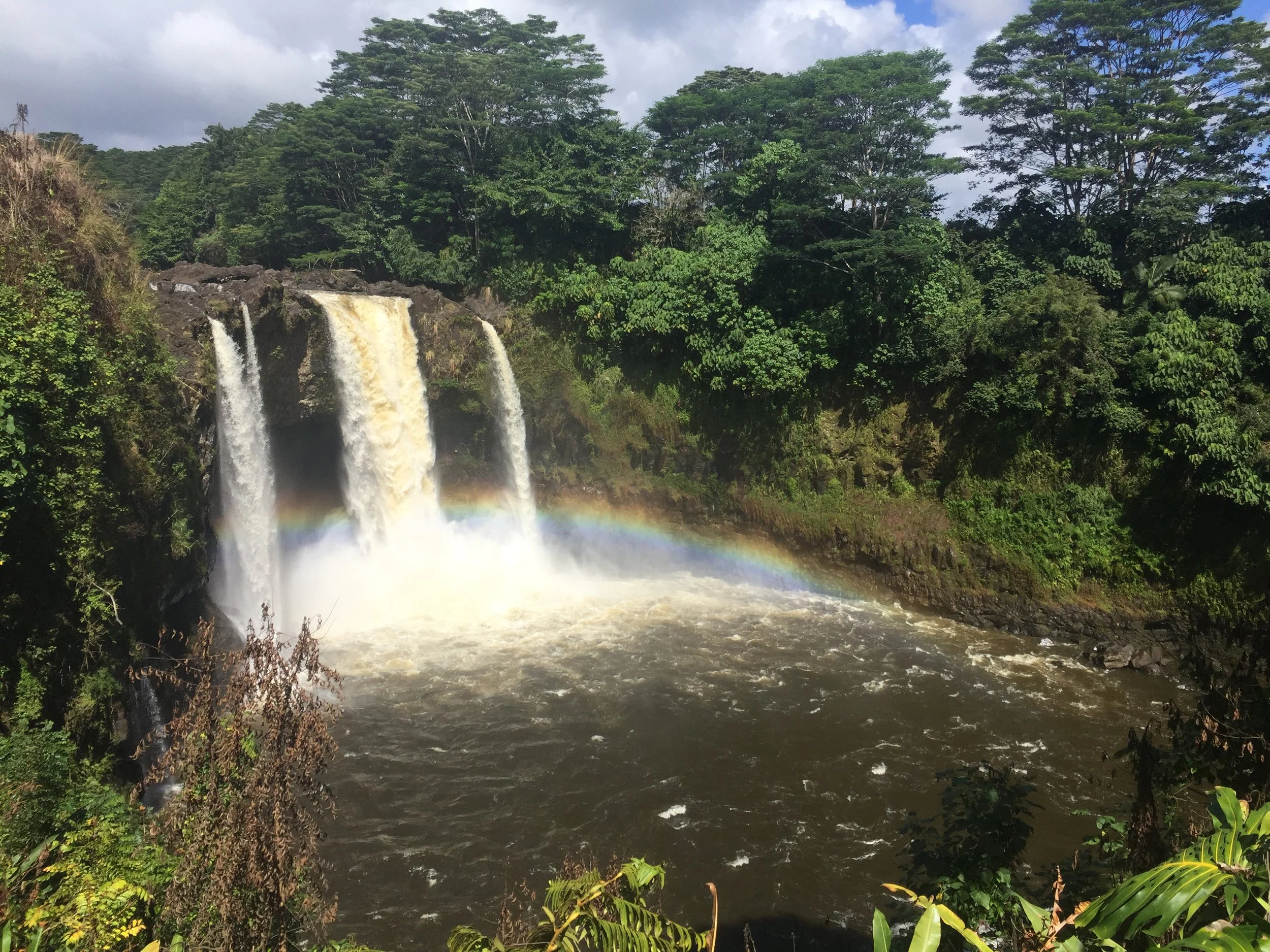 A waterfall with three streams pouring over rocks into a river, surrounded by lush green trees under a partly cloudy sky, with a rainbow visible near the base of the falls.