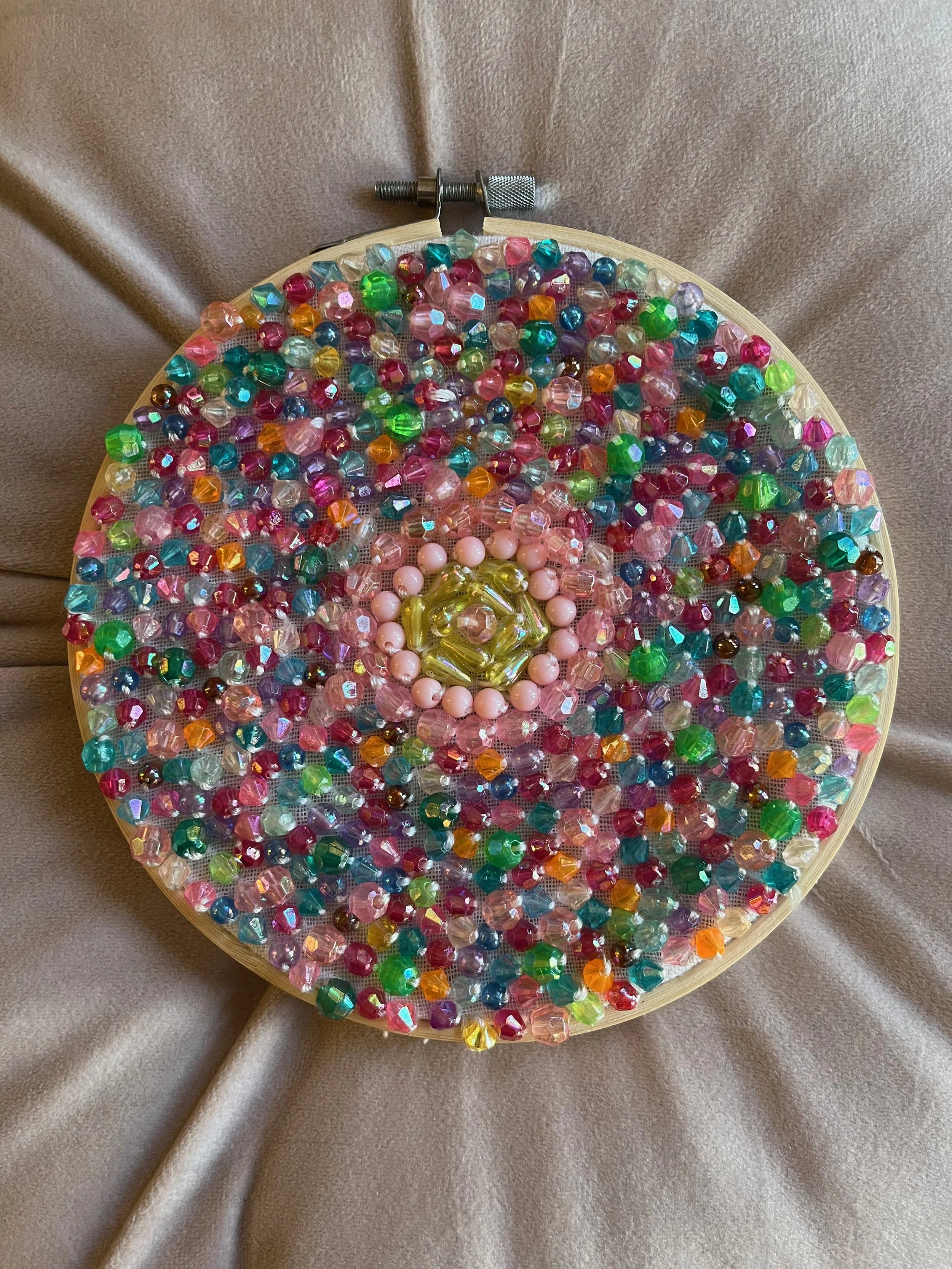 A circular beaded art piece with colorful beads arranged in a spiral pattern, mounted in an embroidery hoop fabric frame.