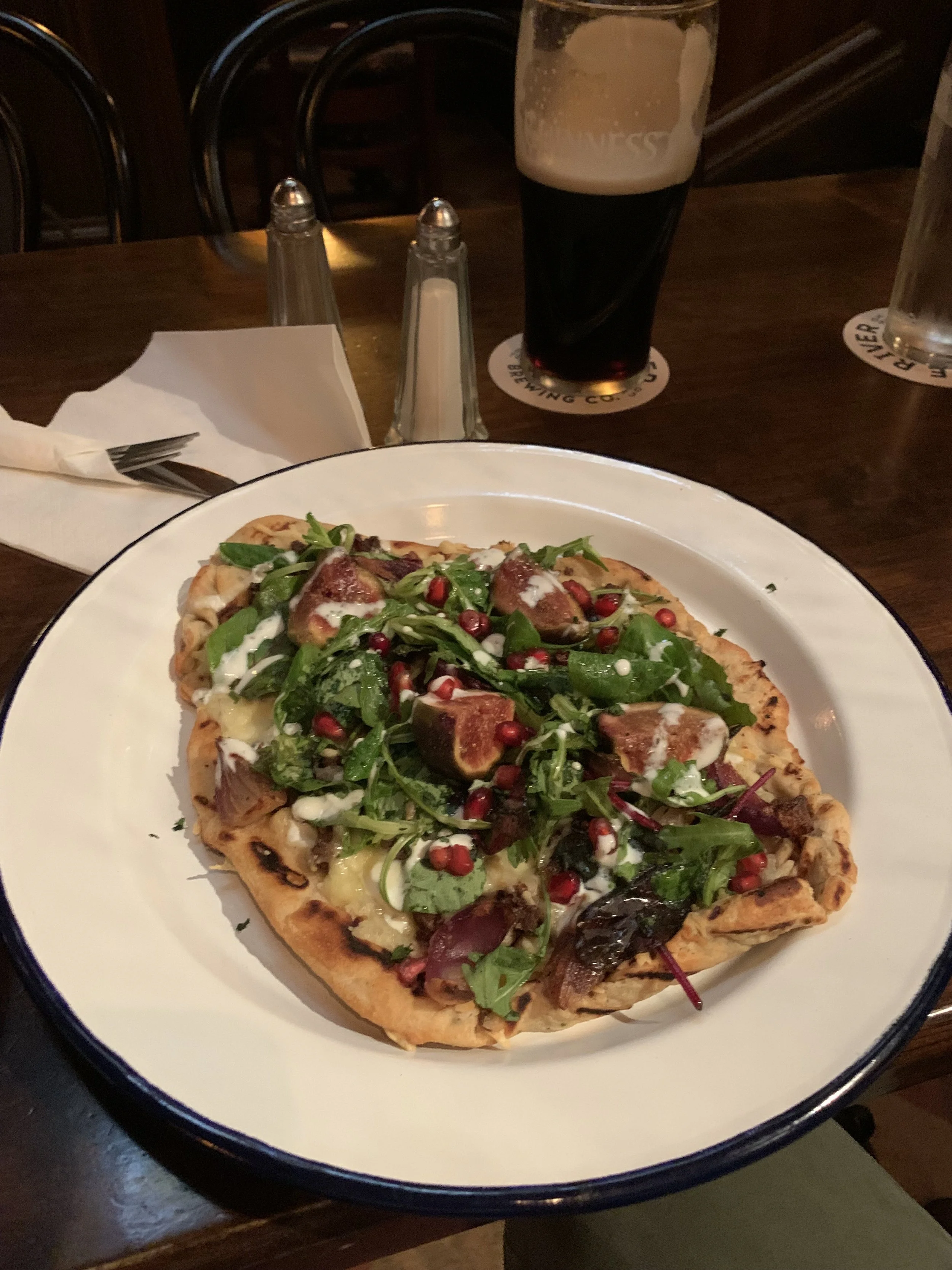 A pizza topped with greens, figs, pomegranate seeds, and drizzled with white sauce on a white plate. In the background, a dark beer in a glass, salt and pepper shakers, and a glass of water on a wooden table.