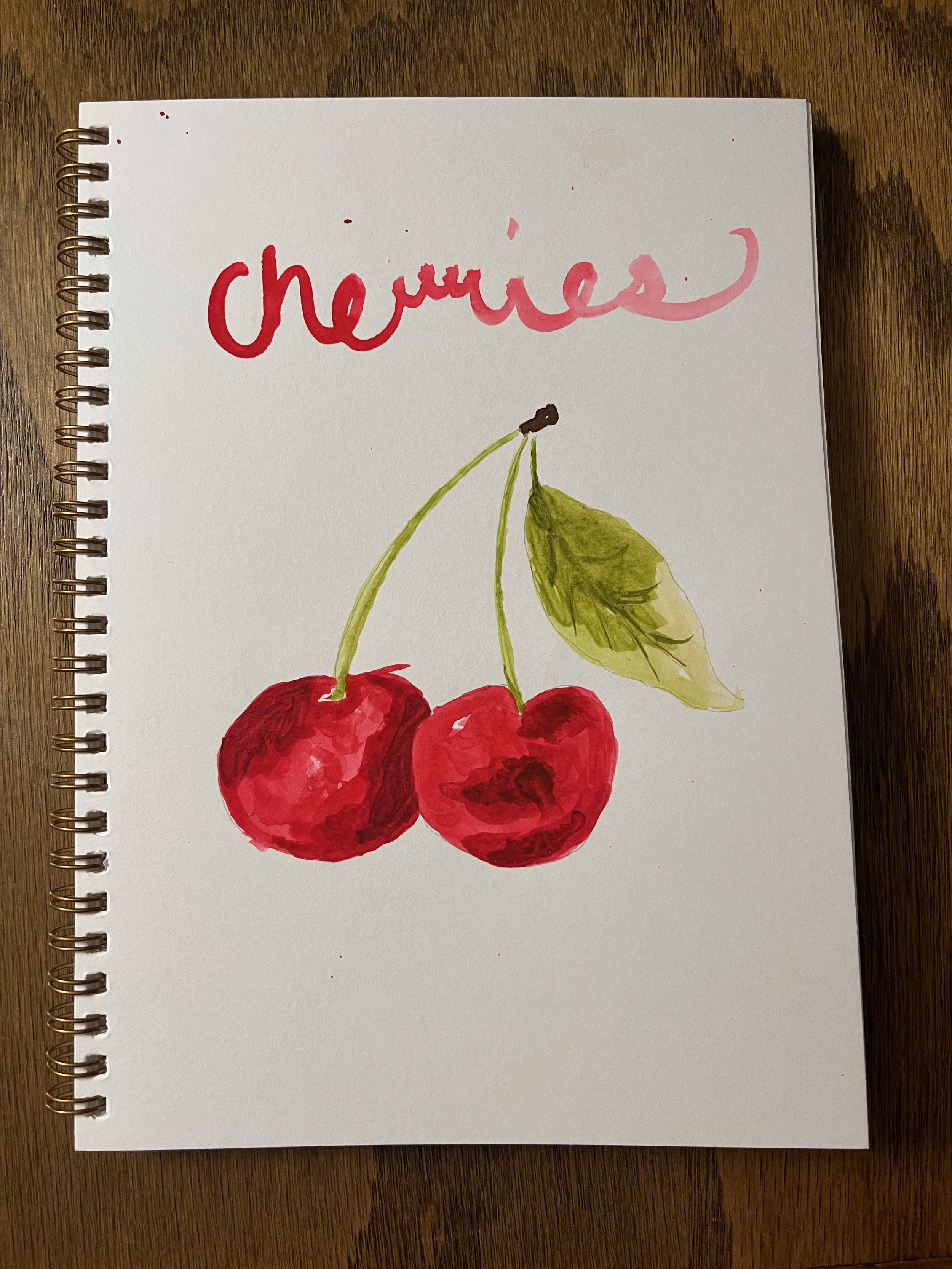 Watercolor drawing of two red cherries with green stems and leaves, with the word 'cherries' written above in pink and red