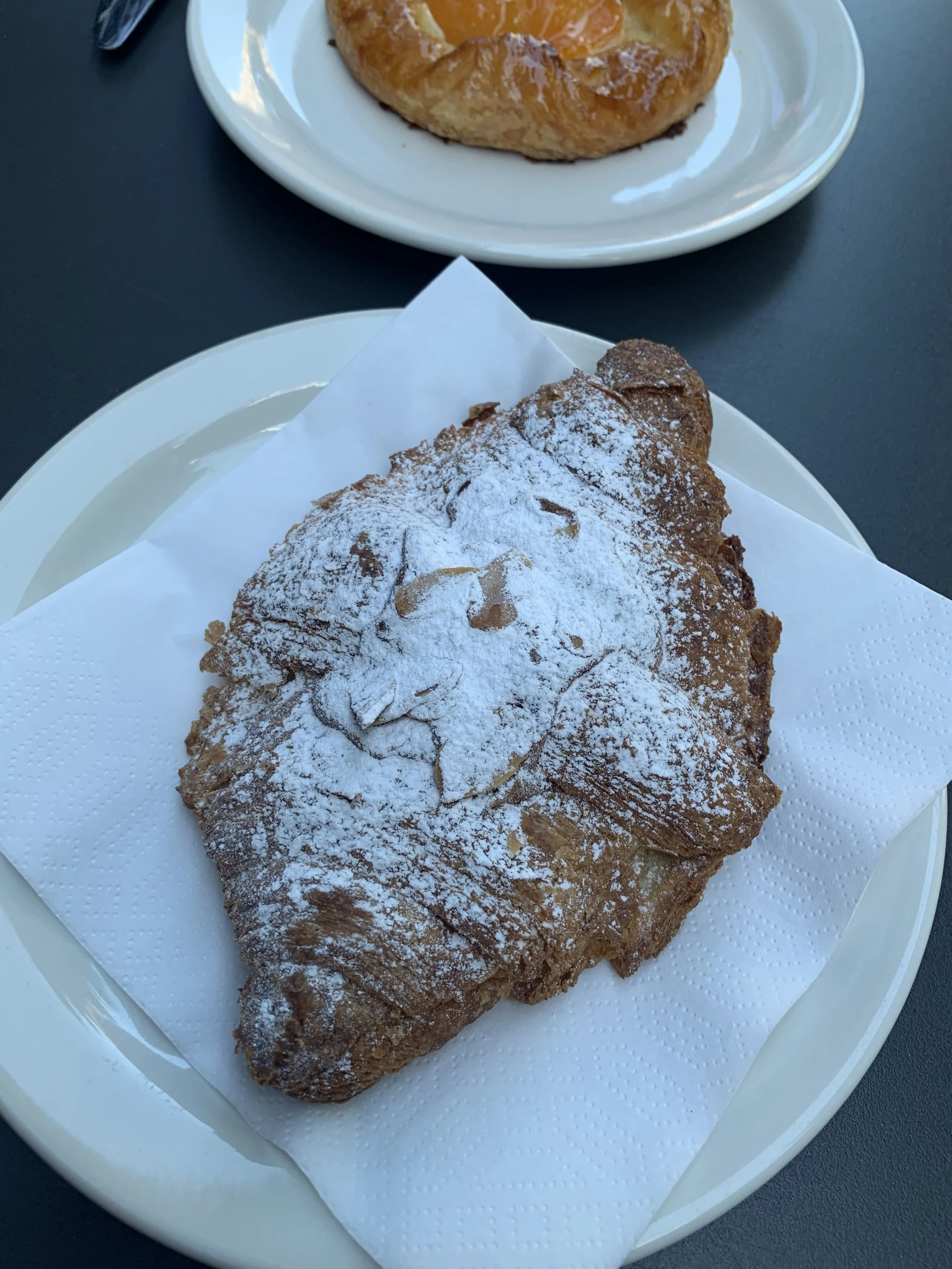 A croissant dusted with powdered sugar on a paper napkin, placed on a white plate. In the background, there is another plate with a pastry or Danish.