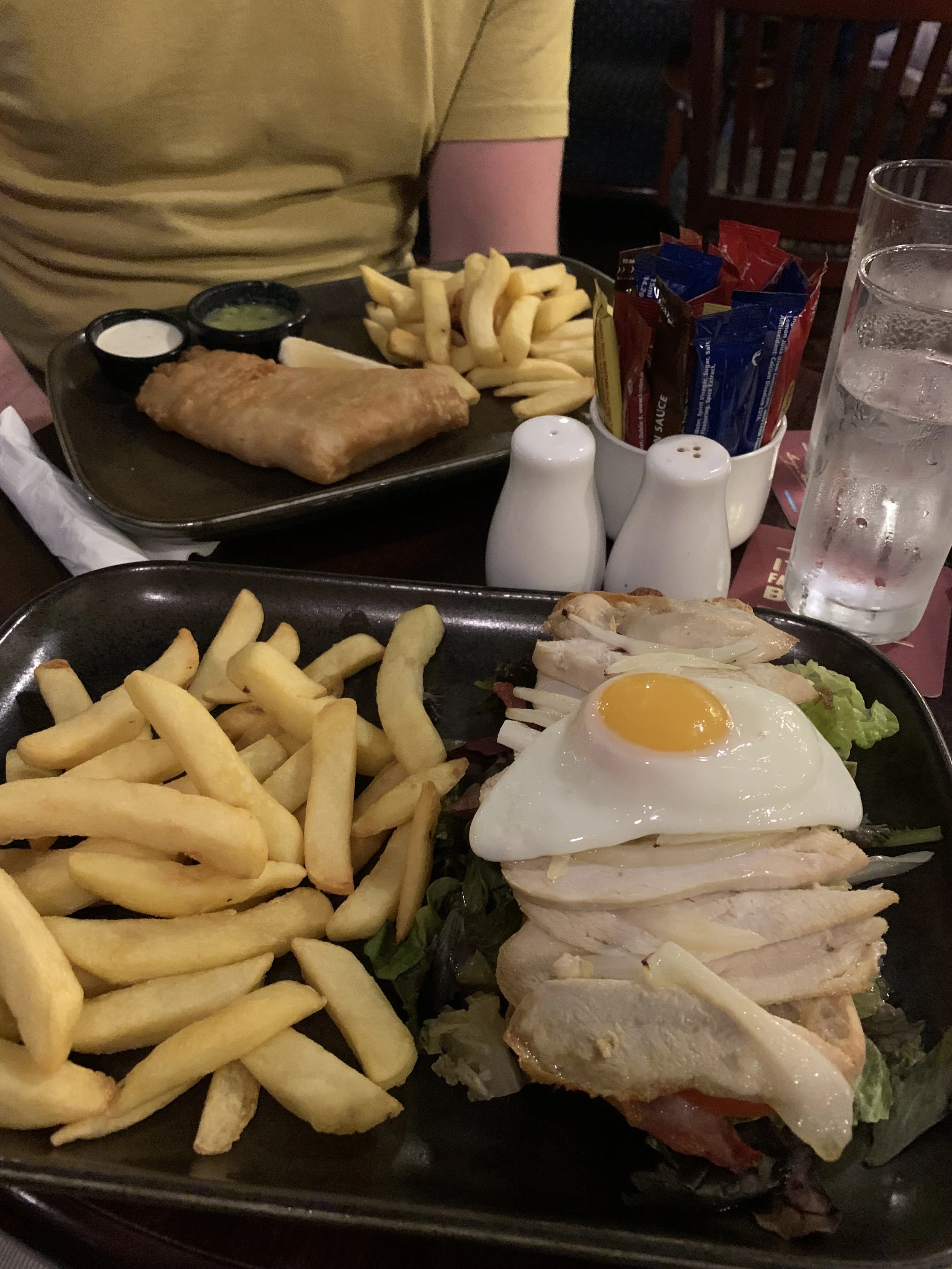 Plates of French fries and chicken salad with a fried egg on top, accompanied by water glasses, salt and pepper shakers, and a plate of fish and chips with tartar sauce on a restaurant table.