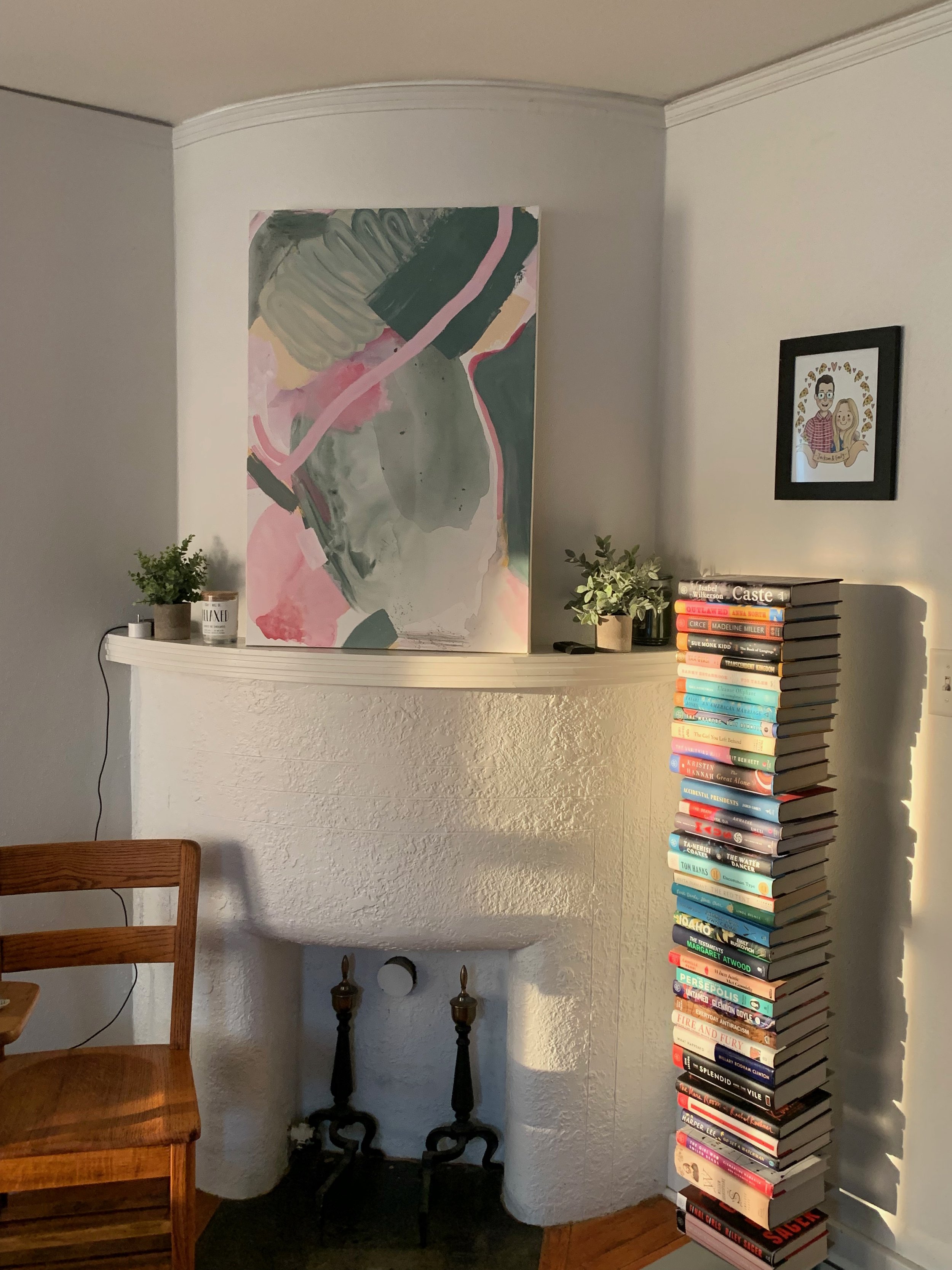 Living room corner with a white textured fireplace, abstract painting above, small potted plants on the mantle, a tall stack of colorful books to the right, and a framed cartoon artwork on the wall. A wooden chair is partially visible on the left.