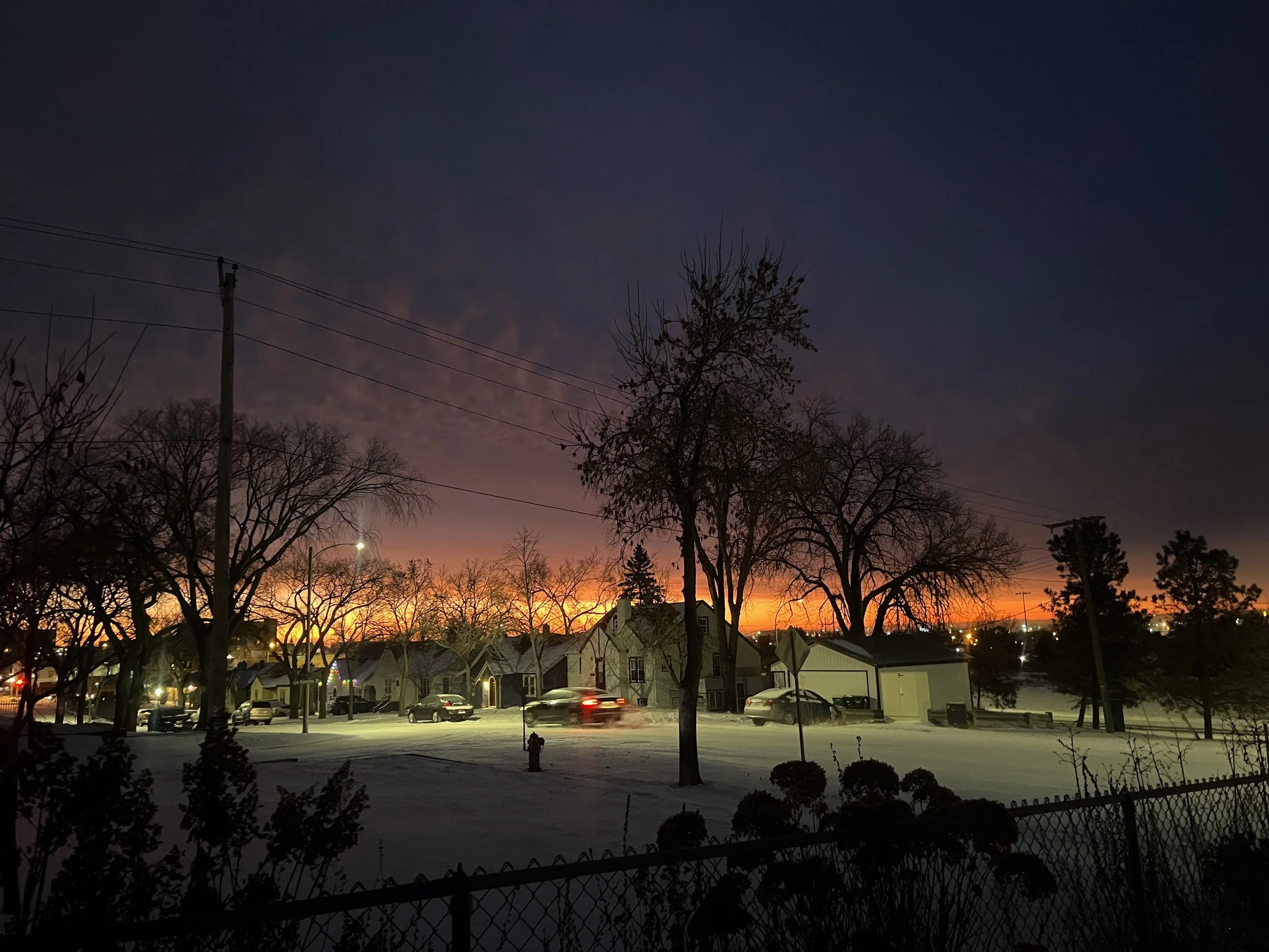 A snowy neighborhood at sunset with silhouetted trees, houses, and cars on a quiet street.