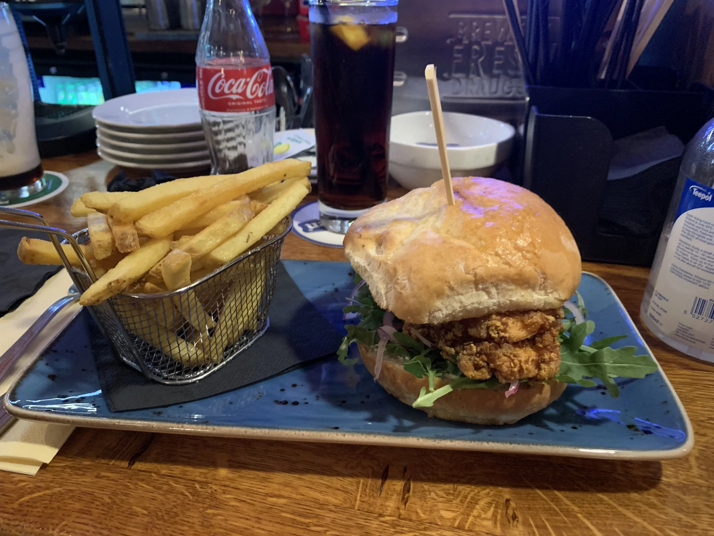 Fried chicken sandwich with lettuce and pickles in a bun, served with a side of French fries in a small metal basket at a restaurant table.