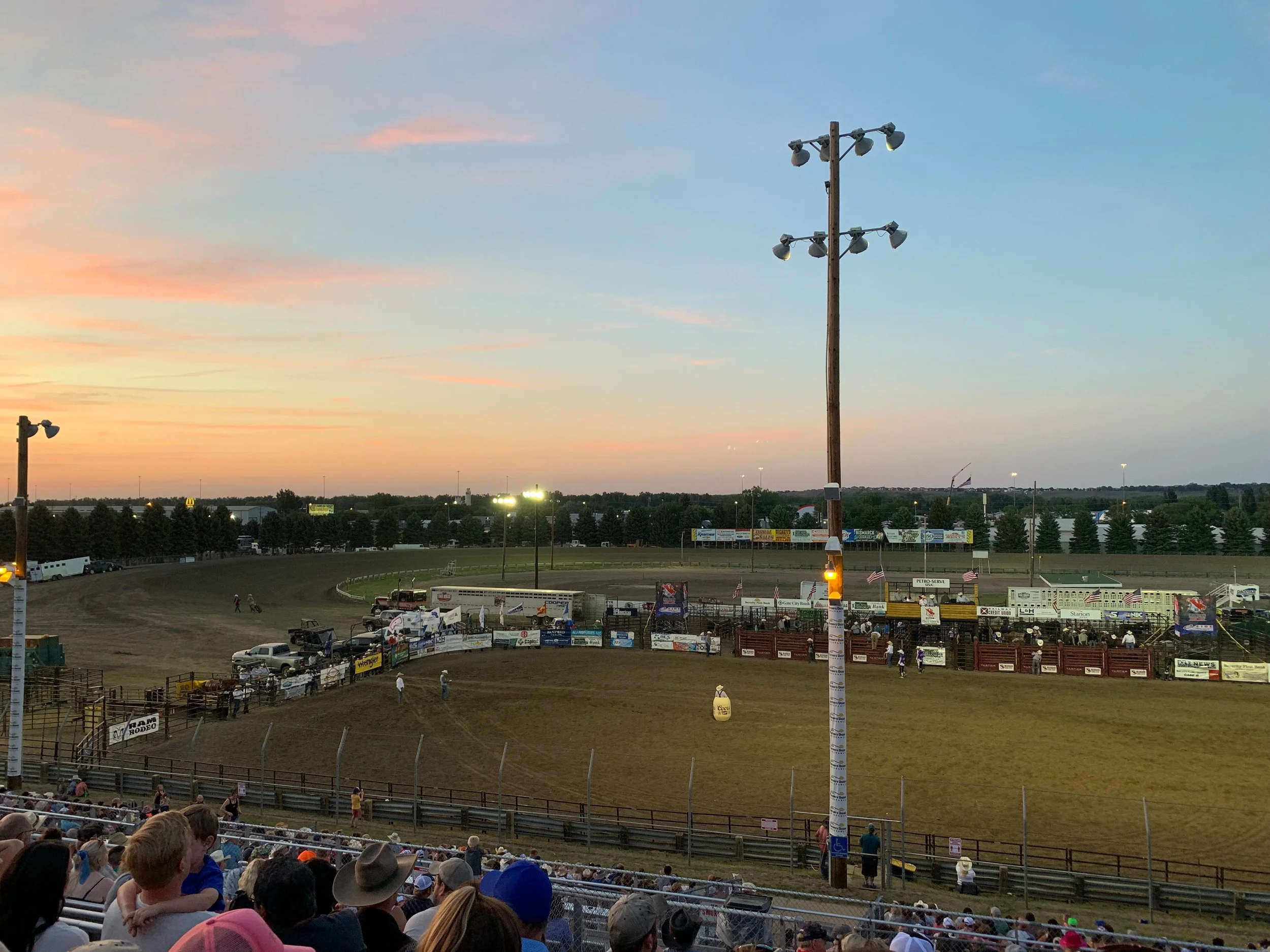 A dirt racetrack at a rodeo with spectators in the stands, evening sky with sunset colors, and stadium lights illuminating the area.