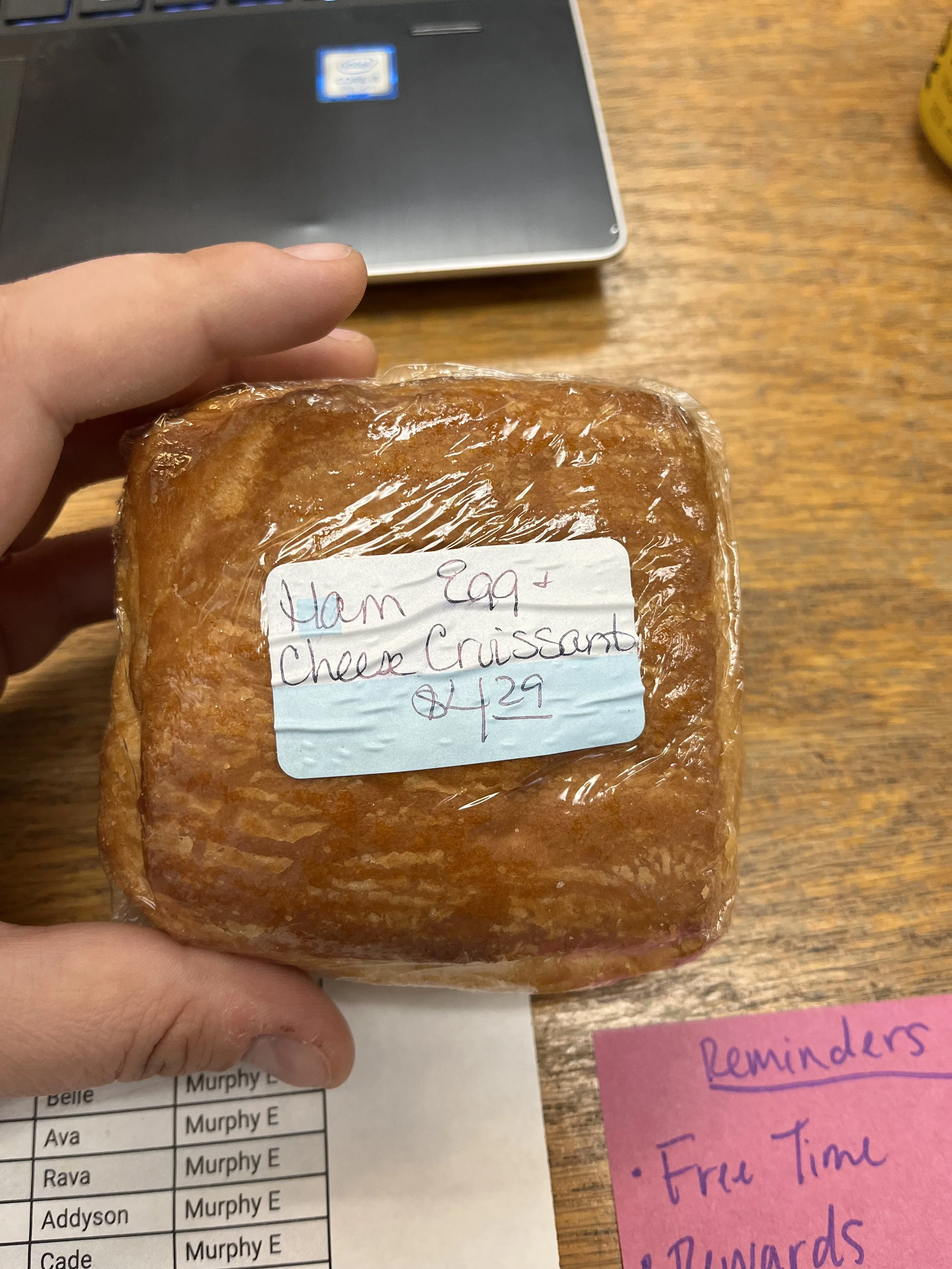 A hand holding a wrapped ham, egg, and cheese croissant sandwich with a handwritten label, on a wooden table near a laptop and pink reminder paper.