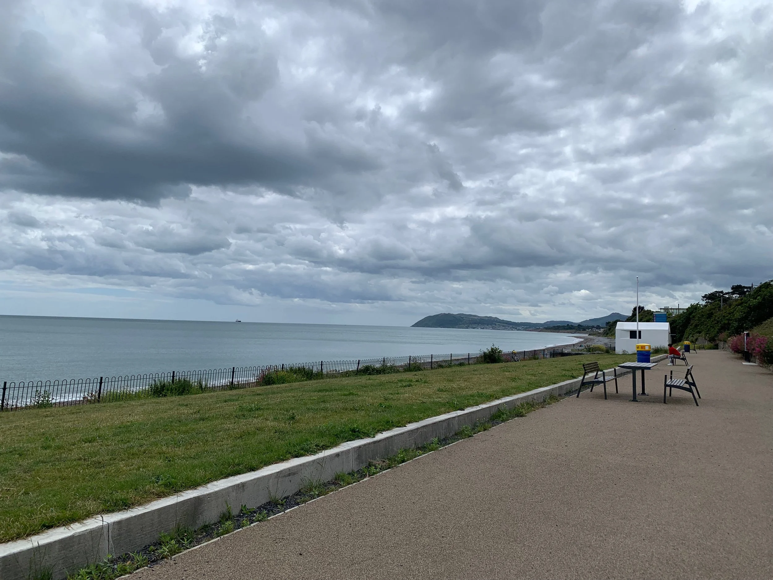 Overcast sky over a coastal promenade with benches, a trash can, a white small building, and a view of the ocean and distant hills.