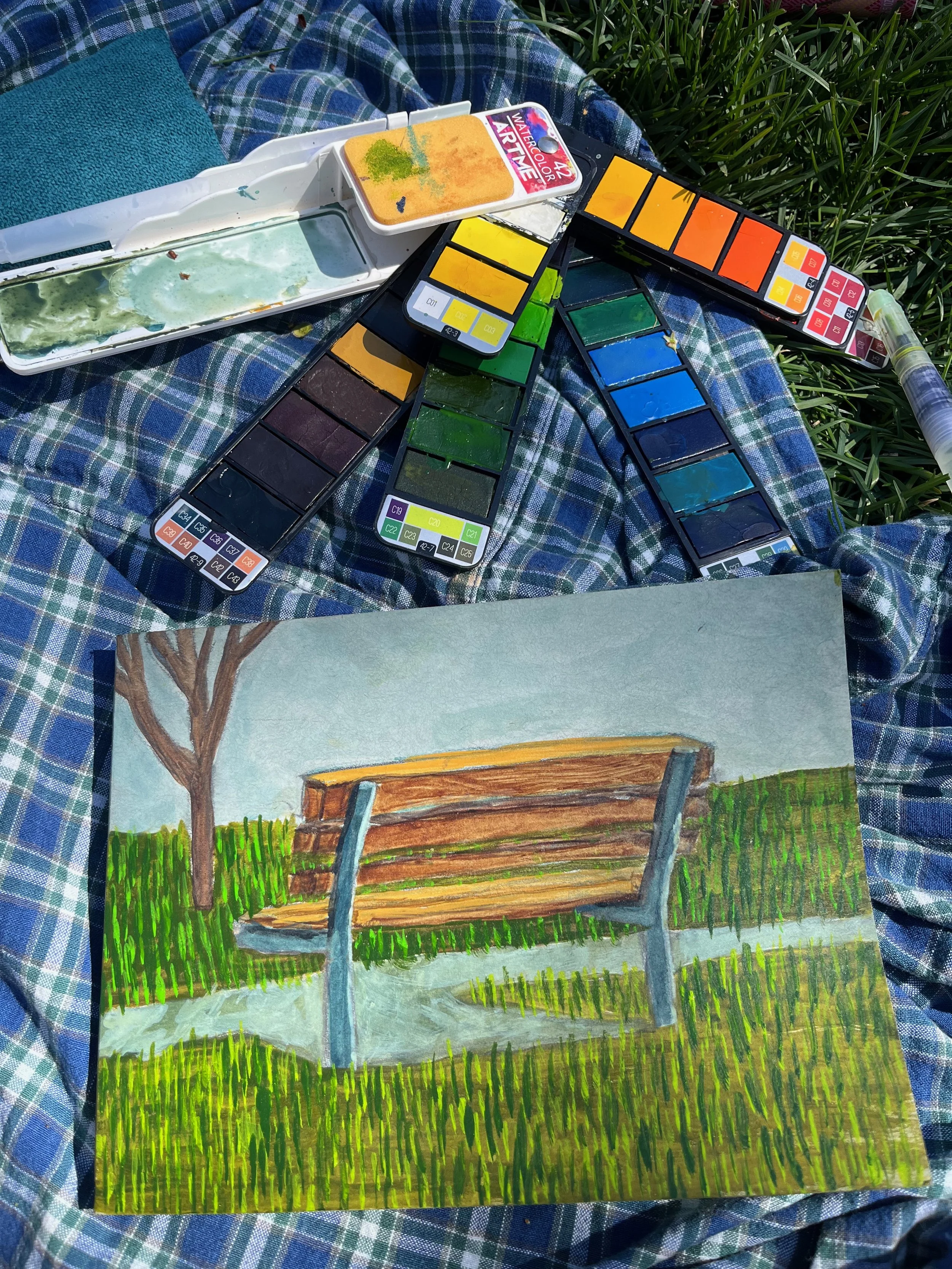 A colorful painting of a park bench with a tree in the background, painted with watercolors on paper. The painting is on a blue plaid fabric, surrounded by watercolor paint palettes and brushes.