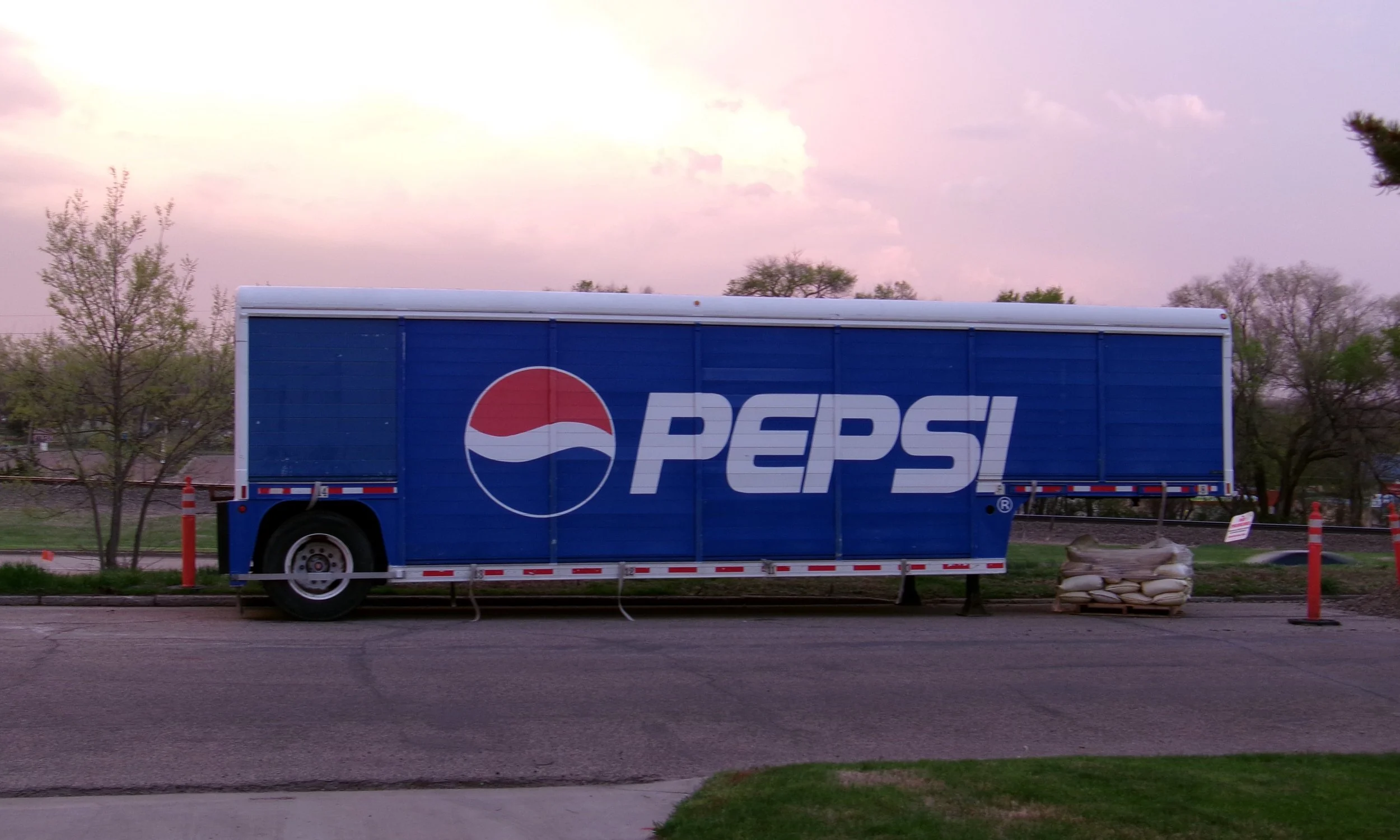A blue Pepsi-branded trailer parked on the side of a street with trees and a cloudy sky in the background.