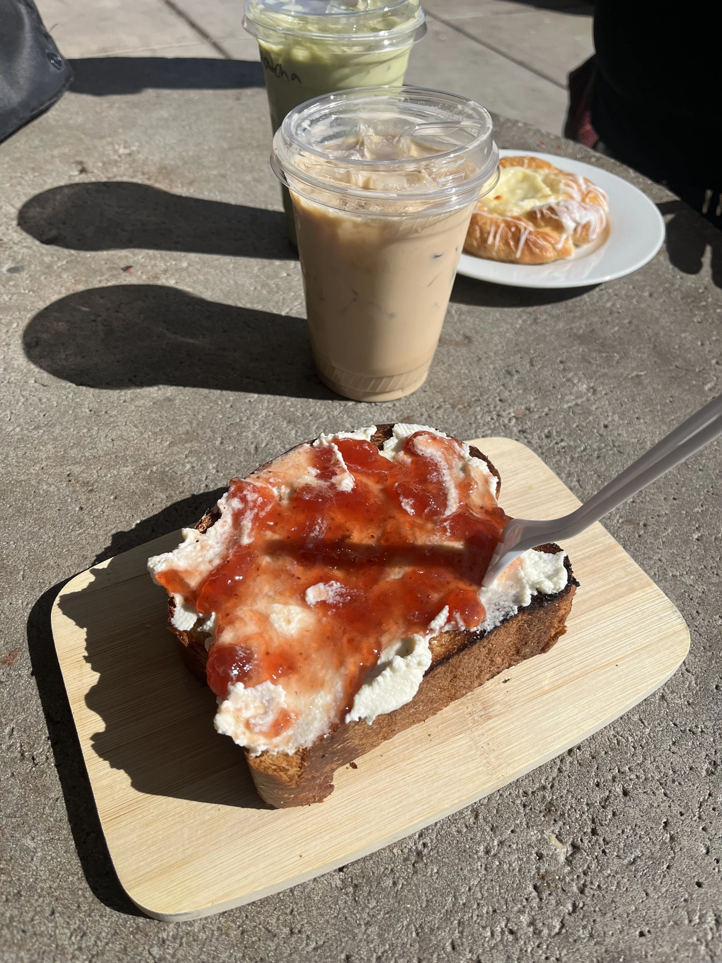 Slice of bread with cream cheese, tomato sauce, and chili sauce, served on a wooden board, with iced coffee in a plastic cup, an iced latte in a plastic cup, and another pastry in the background on a concrete surface.