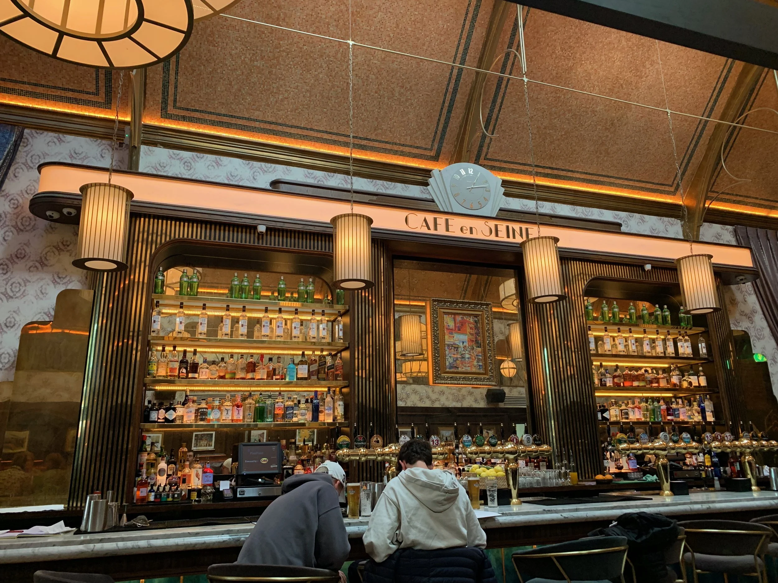 Interior of a bar with two people seated at the counter, shelves filled with bottles of alcohol, and hanging lamps, in a restaurant named 'Café en Seine'.