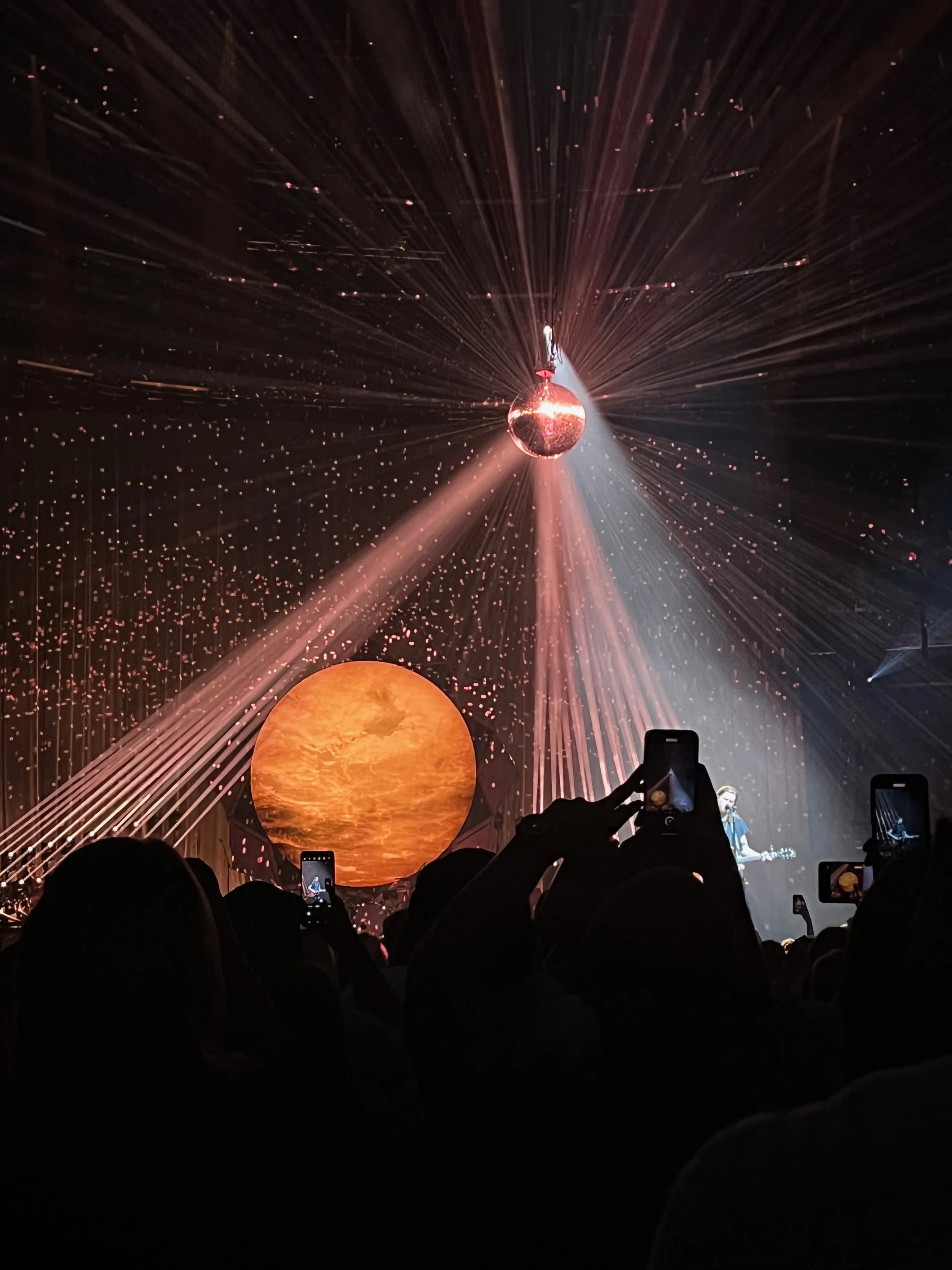 A concert scene featuring a singer playing guitar on stage with a large orange sun visual in the background, beams of light and sparkles, audience members taking photos.