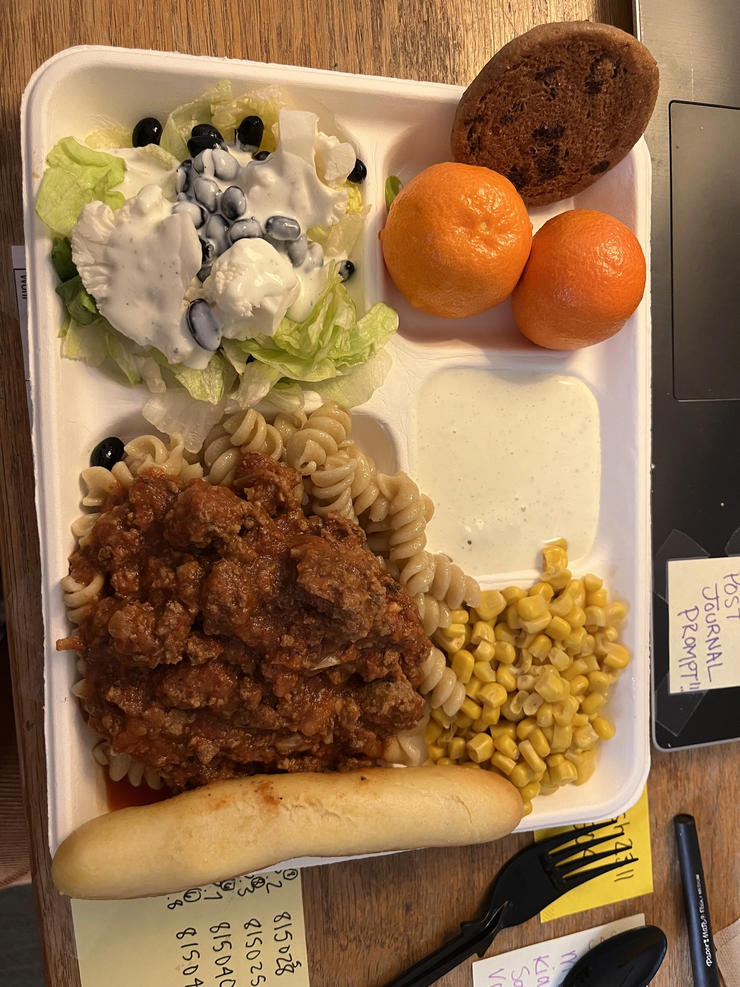 Compartmentalized meal tray with salad topped with dressing, three tangerines, a chocolate chip cookie, pasta with meat sauce, creamed corn, a bread roll, and ranch dressing.