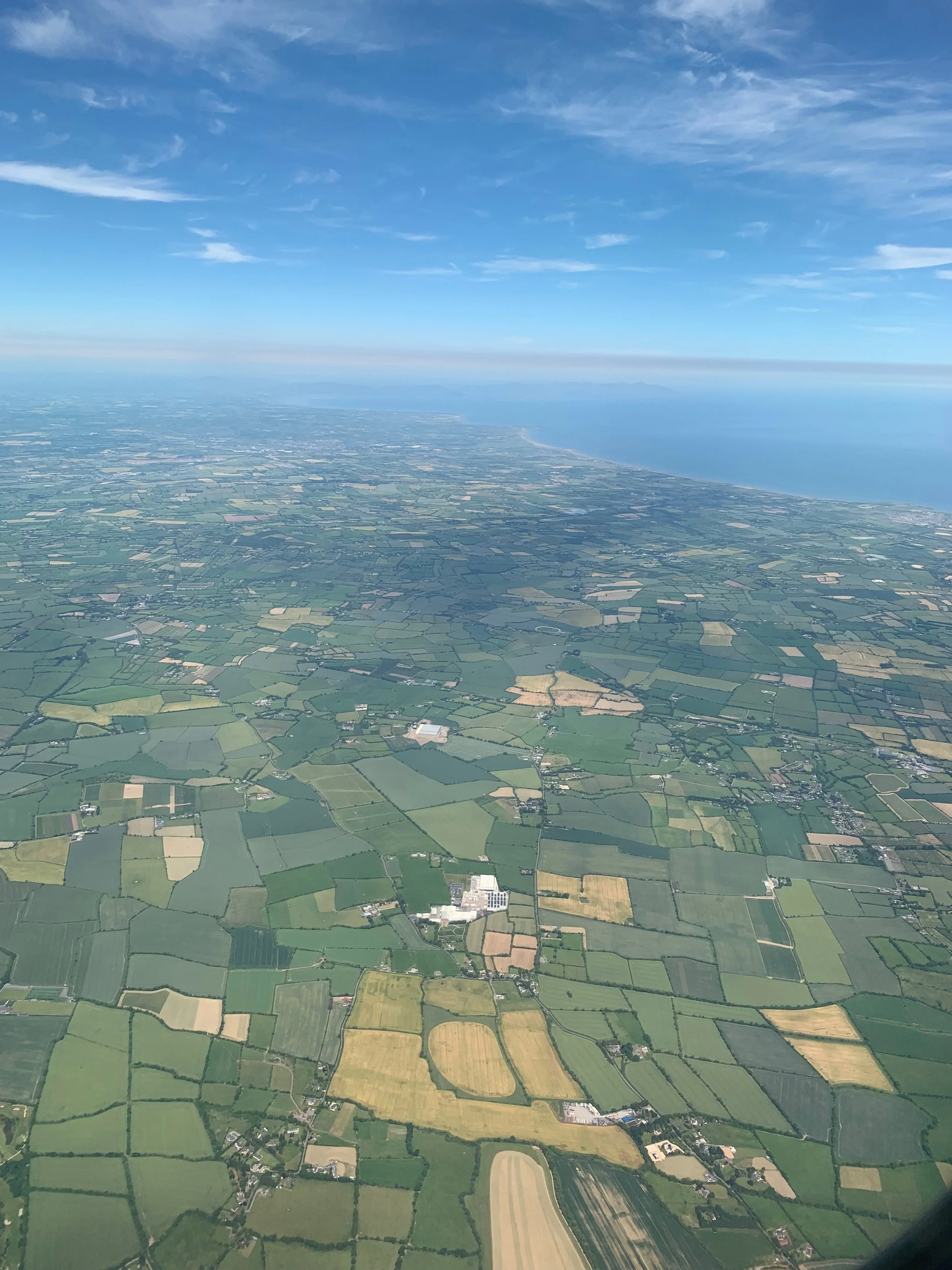 Aerial view of a patchwork of green and yellow farmland with a coastline in the distance under a partly cloudy sky.