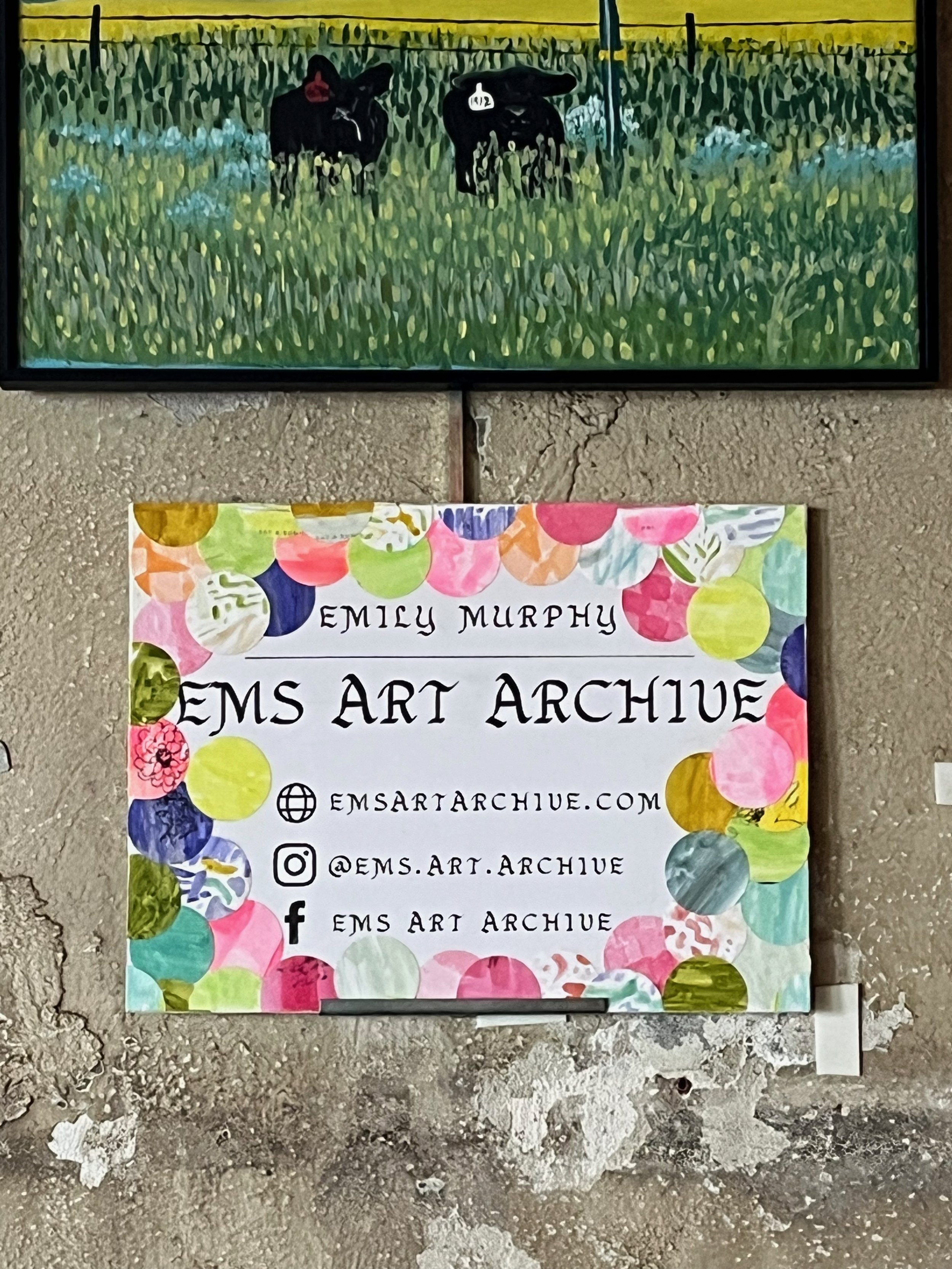 Colorful sign with floral border displaying contact information for EMS Art Archive, including website, Instagram, and Facebook handles, placed on a rough concrete wall below a landscape painting.