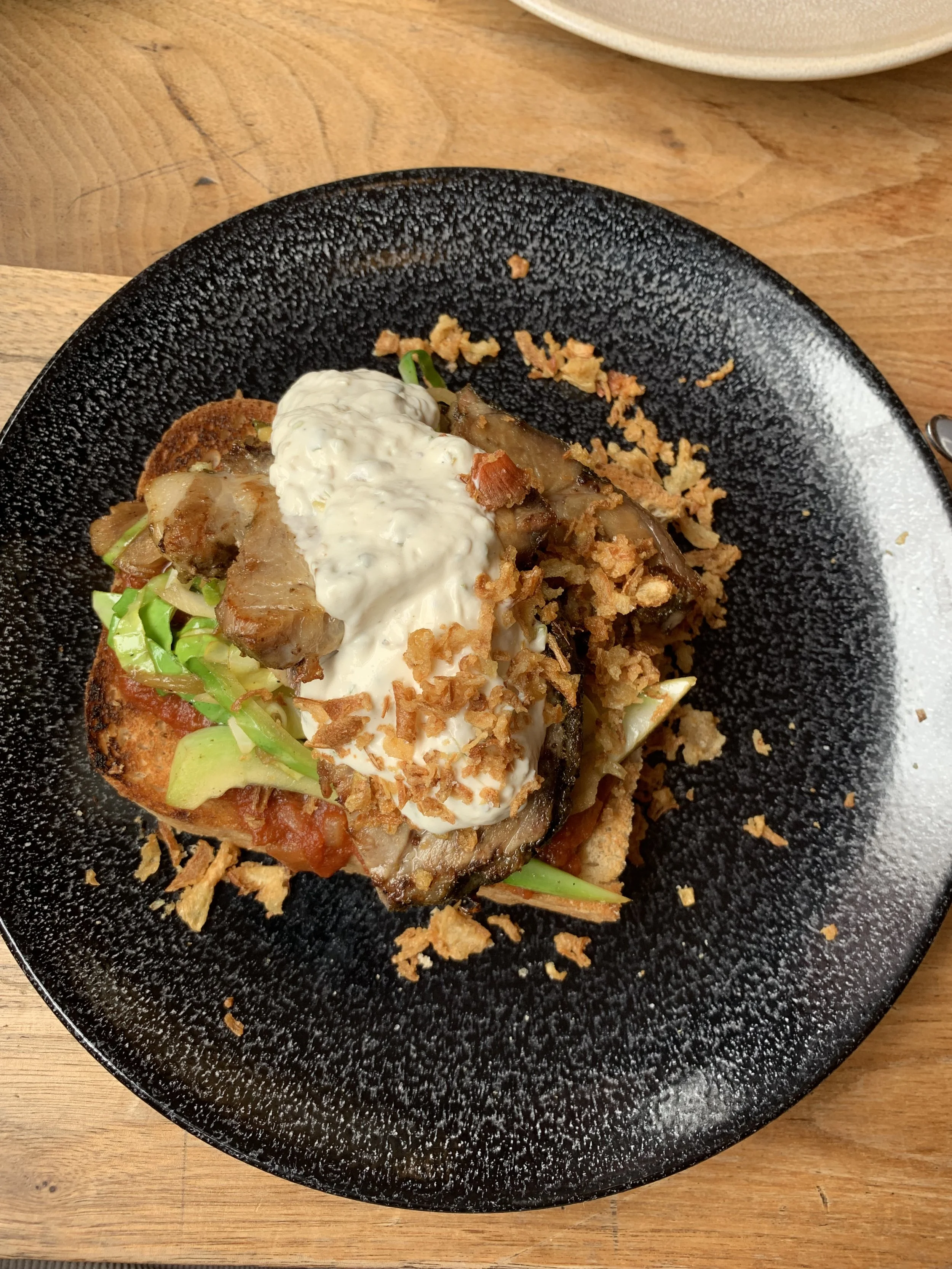 A plated dish featuring a generous portion of pork belly topped with sour cream and crispy onions, served with vegetables on a black plate.