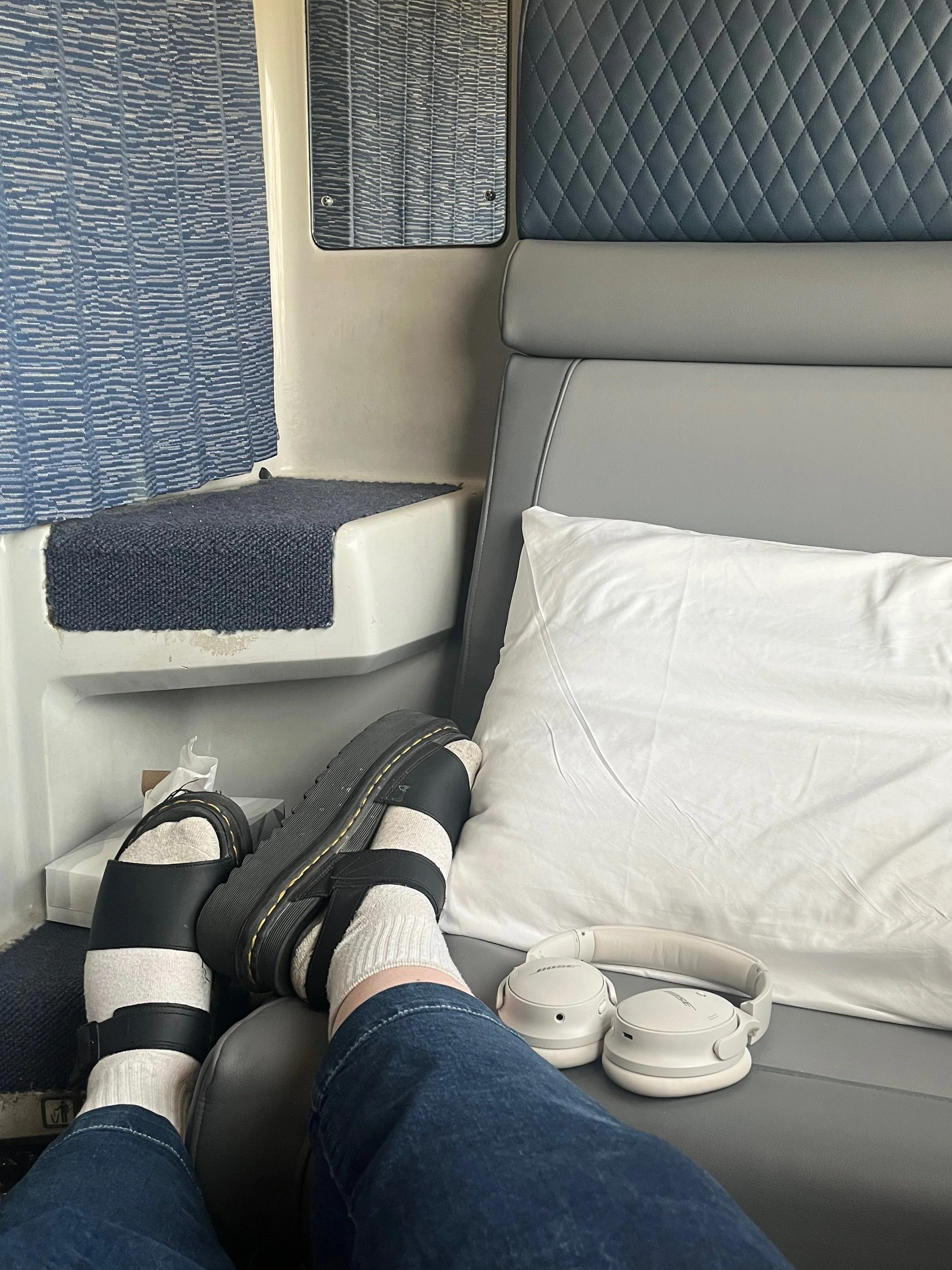 View of a person's legs wearing sandals and white socks, resting on a gray airplane seat with a white pillow, headphones, a tissue box, and a window with blue curtains.