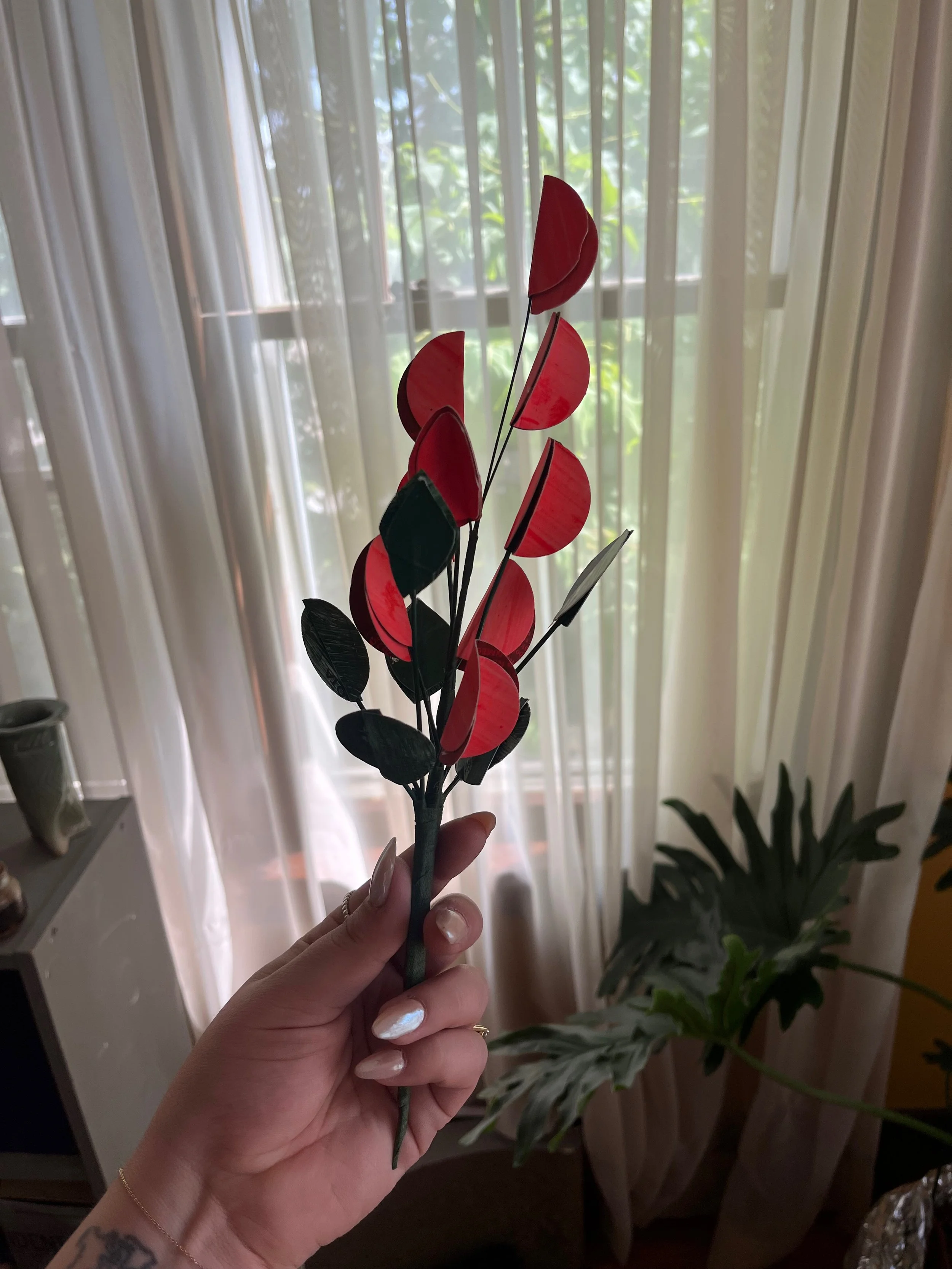 A hand holding a bouquet of artificial red and black leaves with a window and curtains in the background.