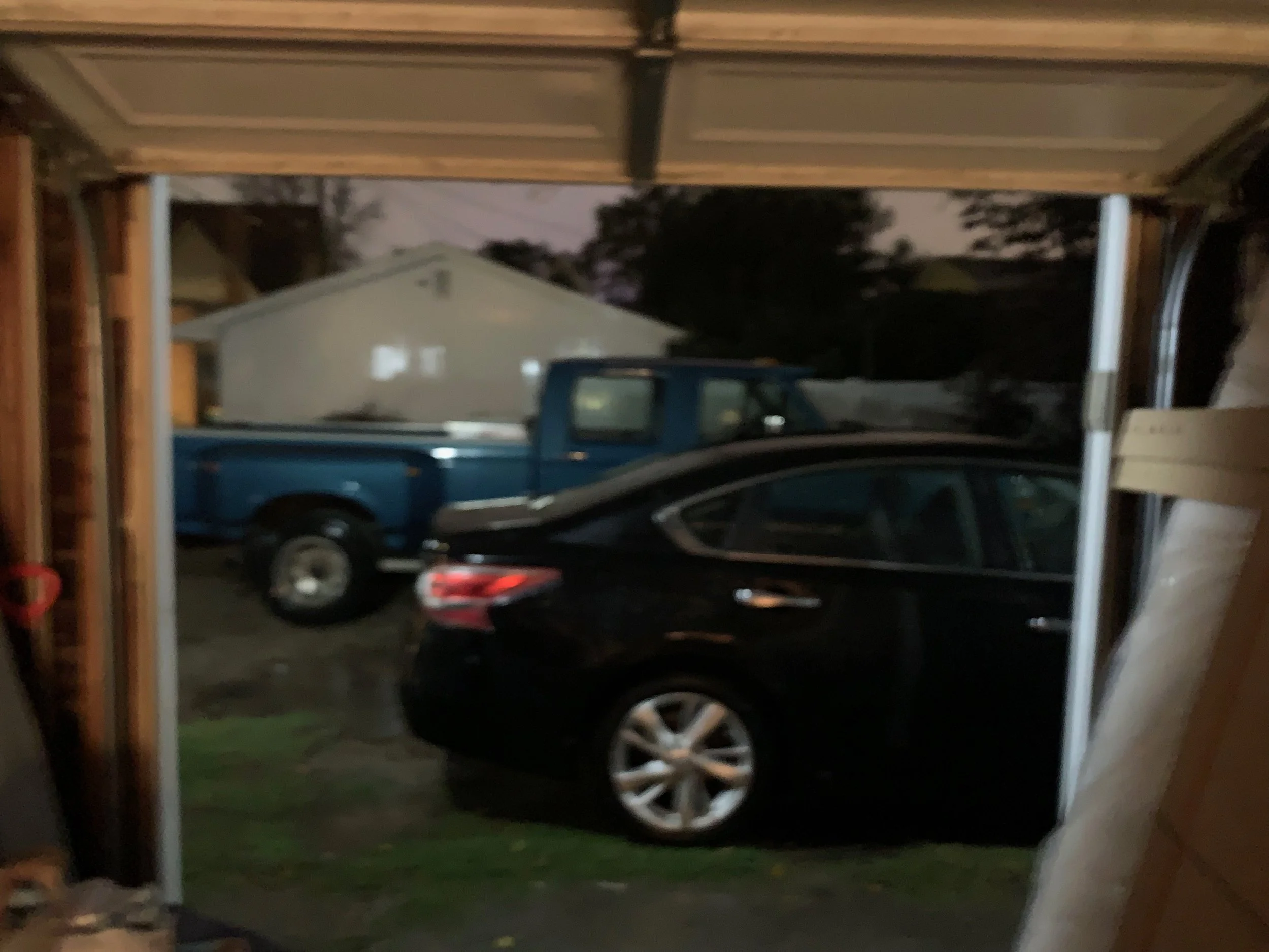 A view from inside a garage looking out at a driveway with a black car and a blue pickup truck parked outside, during dusk or early evening.