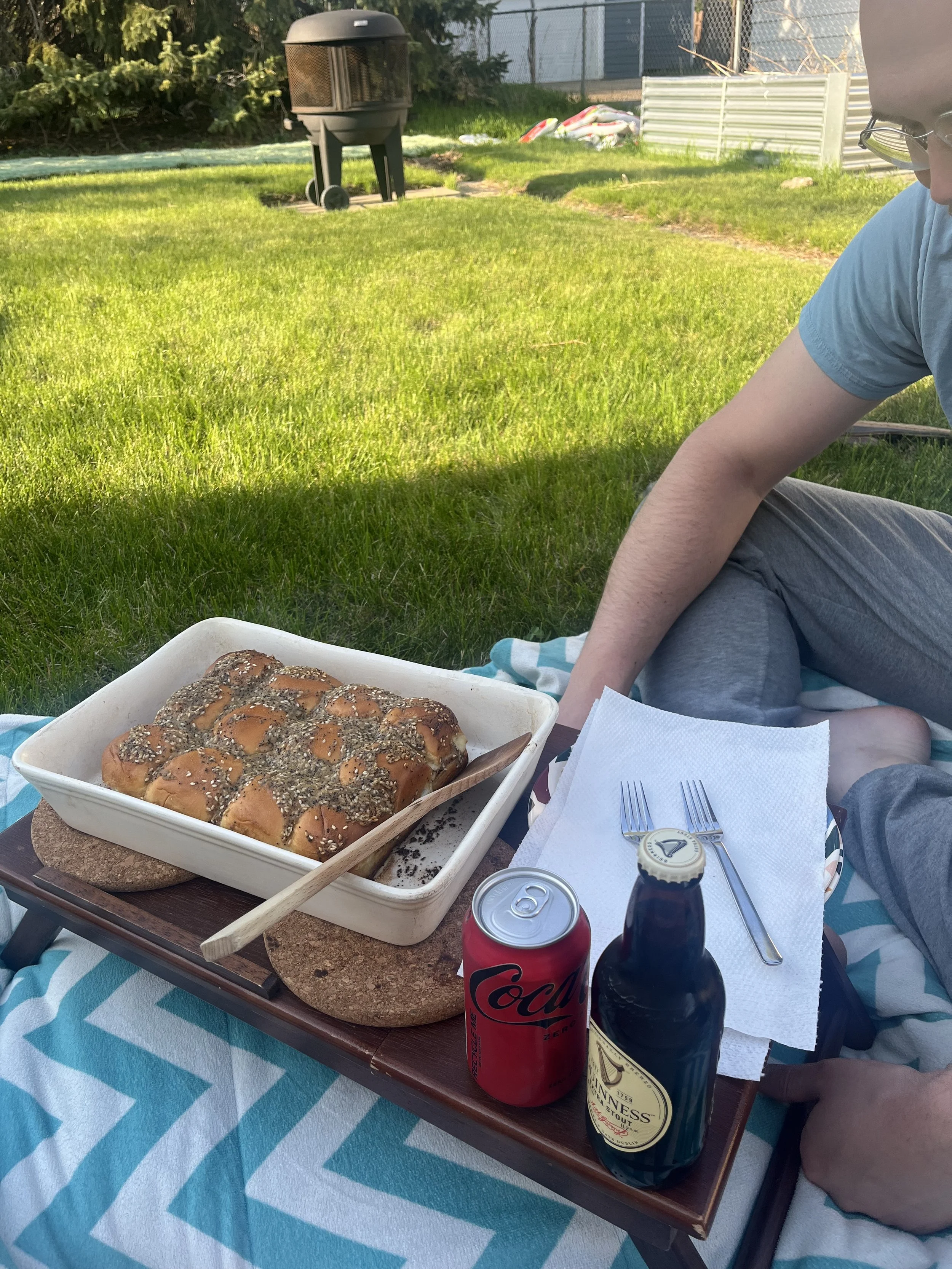Picnic setup on a blanket in a backyard with a tray of freshly baked bread topped with seeds, two cans of soda, and a bottle of Guinness beer. A person sitting nearby is visible.