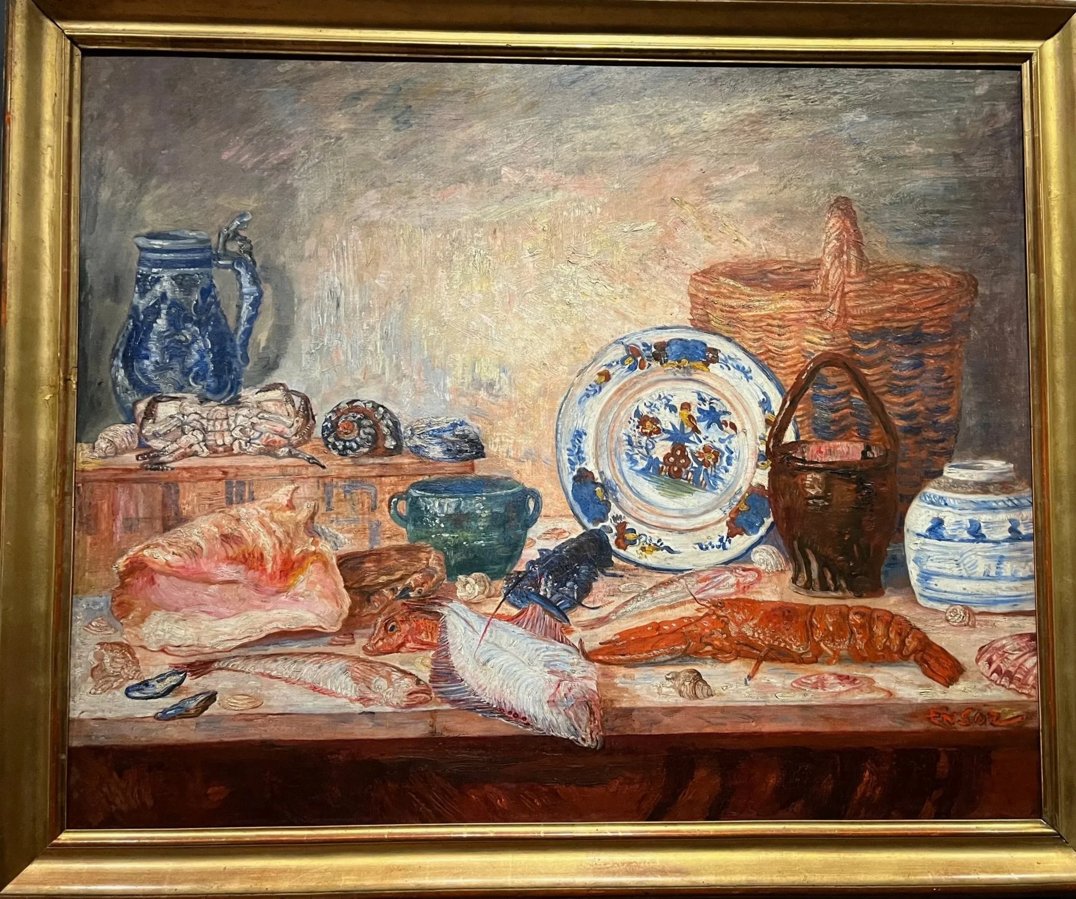 Still life painting of various seafood and kitchenware on a table, including fish, crab, shells, plates, bowls, a basket, and decorative items.