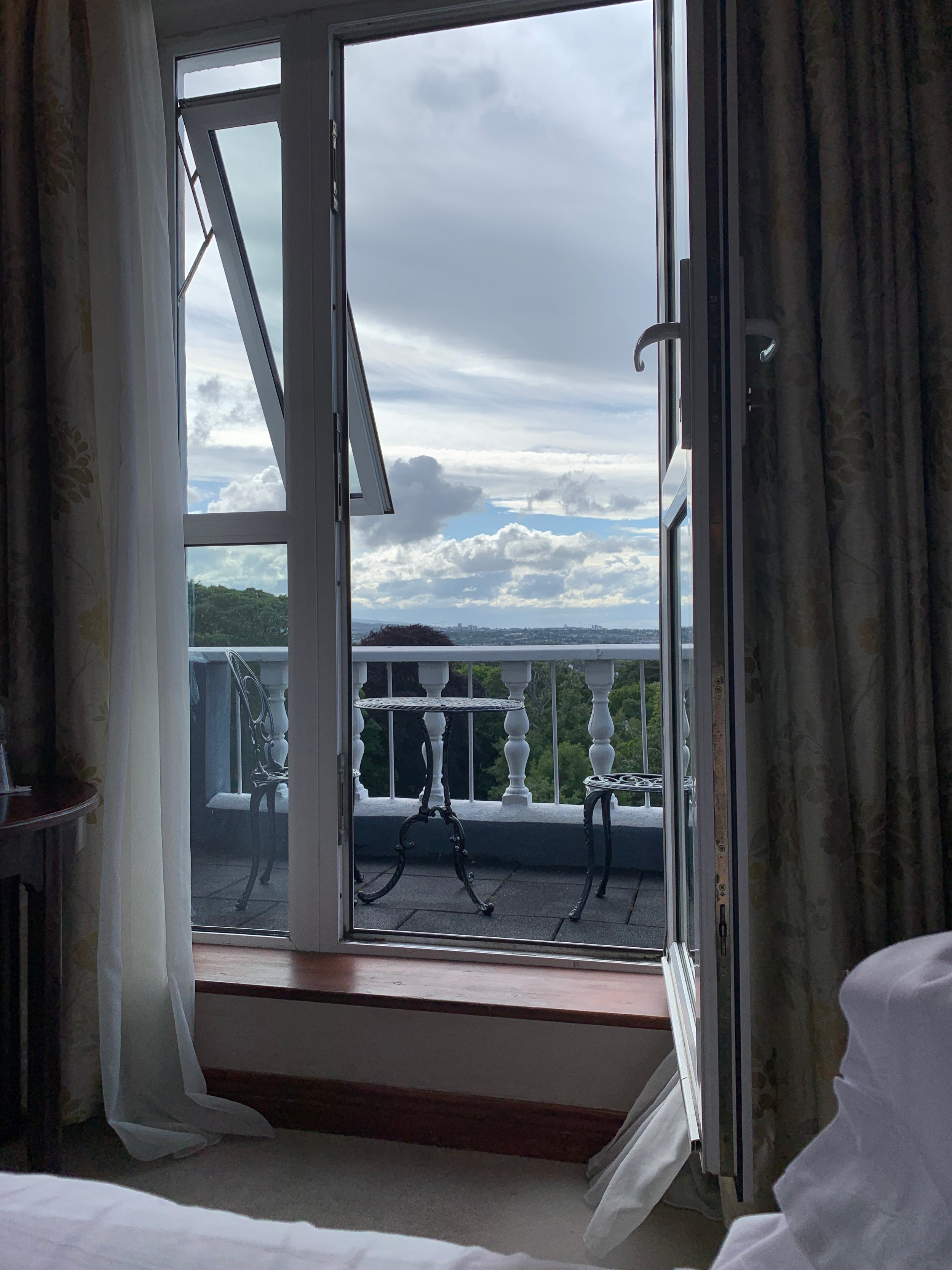Open balcony door looking out from a room with curtains, revealing a small outdoor balcony with a table and two chairs, and a view of cloudy sky and trees in the distance.