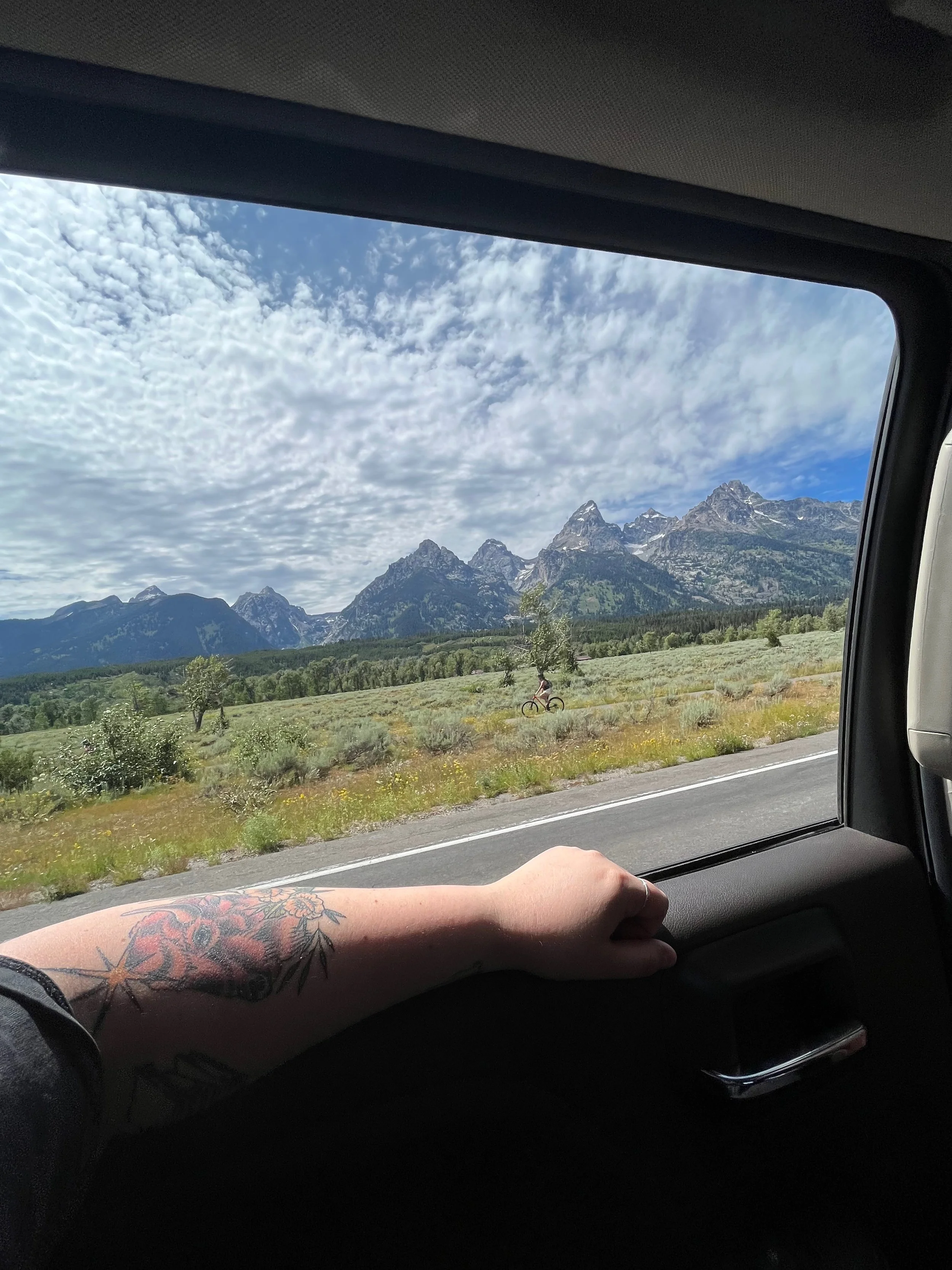 View of mountain range through the car window, with a person's tattooed arm resting on the window ledge, and a cyclist riding on the road outside.