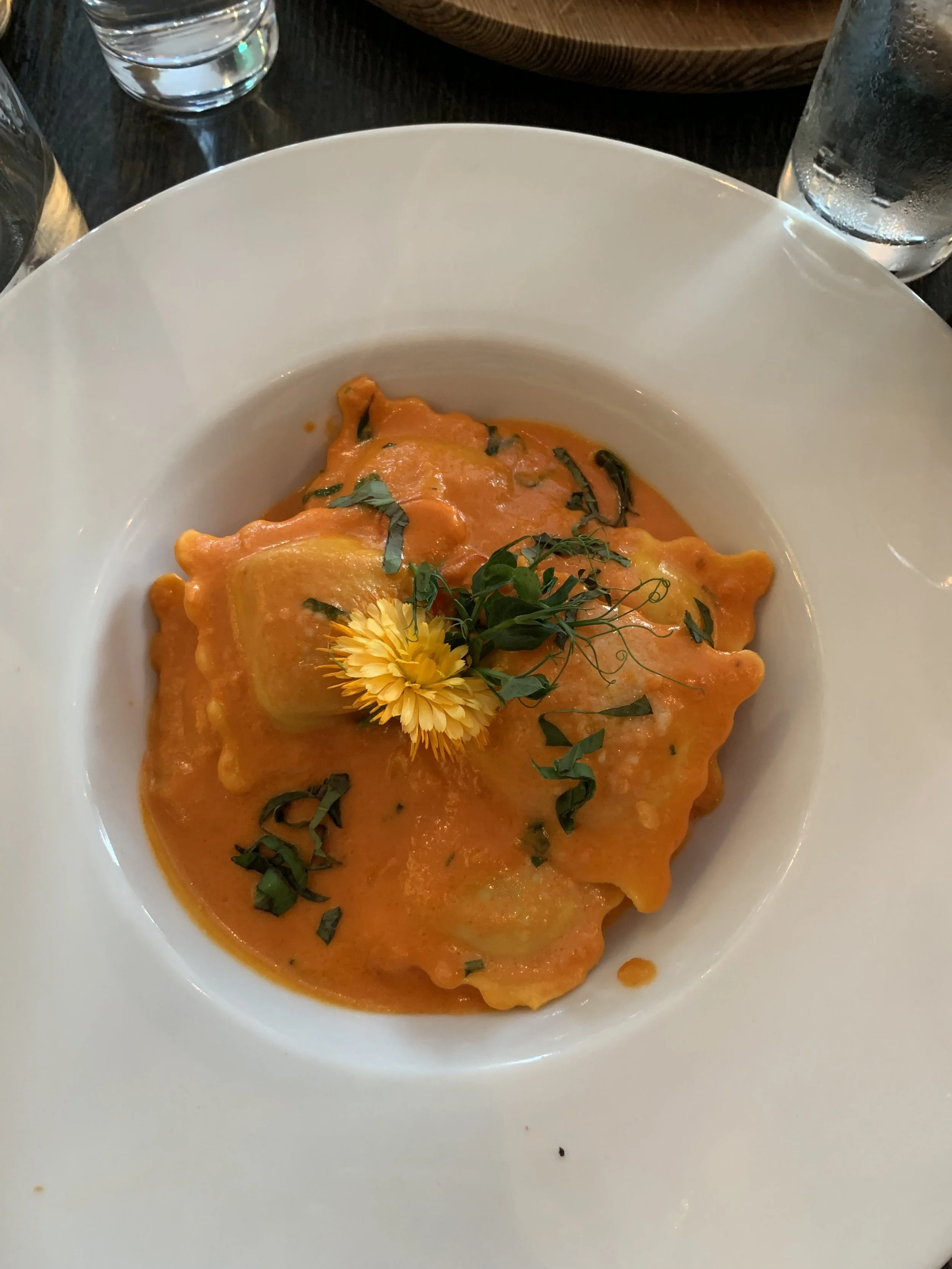 Plate of ravioli covered in orange sauce, garnished with a yellow edible flower and chopped herbs, on a white wide-rimmed plate.