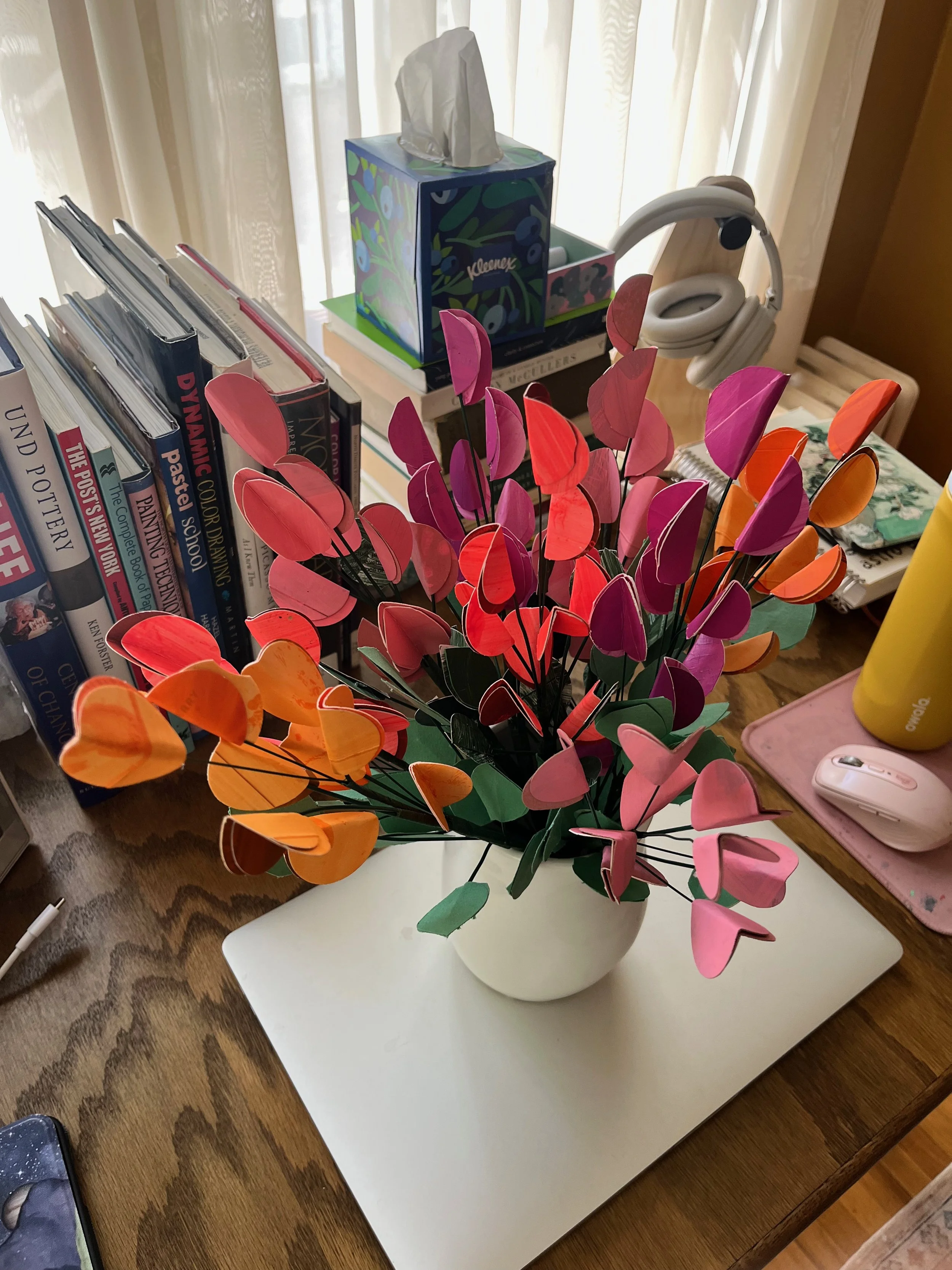 Colorful paper heart-shaped flowers in a white vase on a wooden desk, with books, tissue box, headphones, and a yellow tumbler in the background.