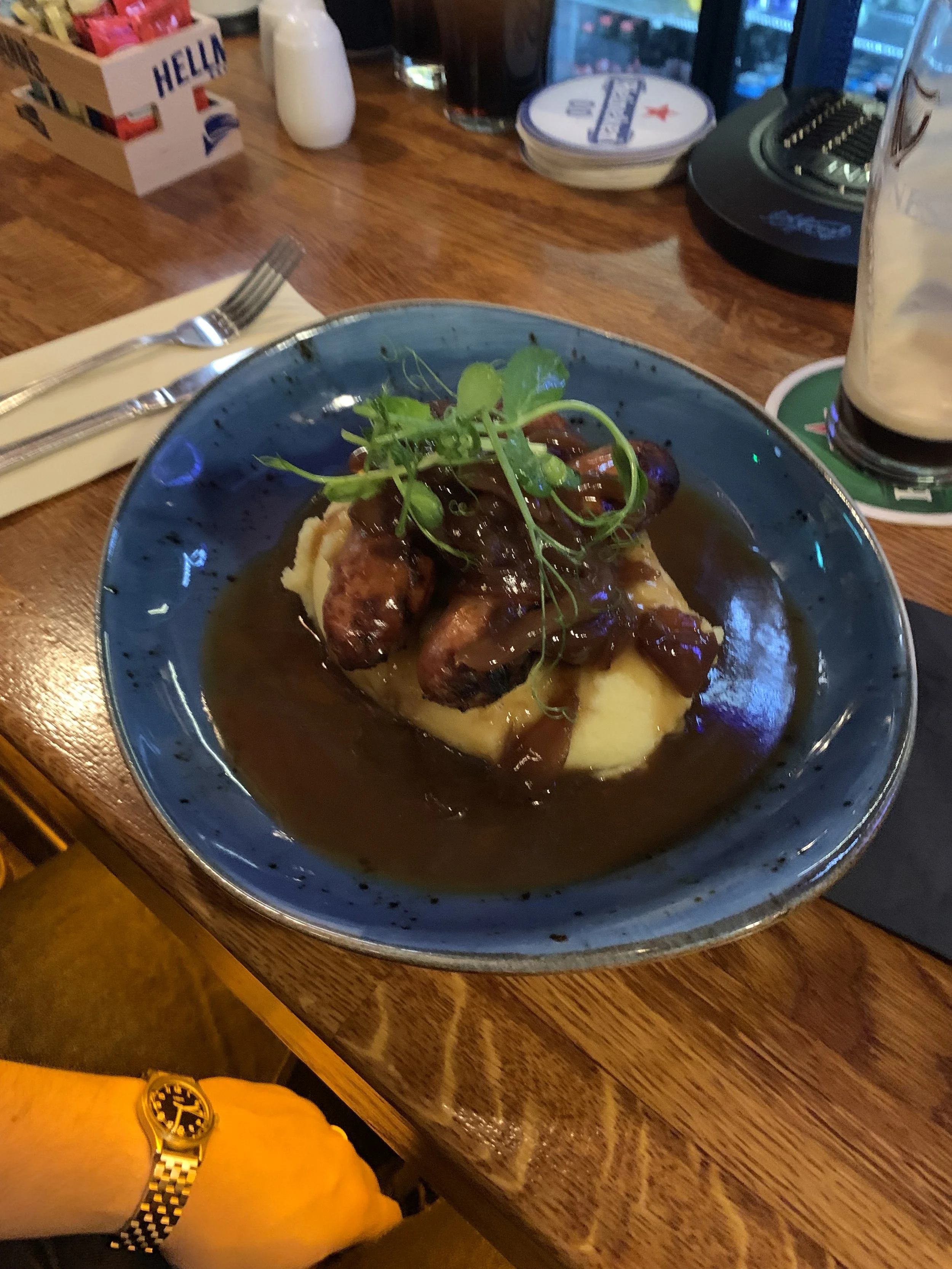 A plate of mashed potatoes topped with mini sausages, brown gravy, and garnished with green herbs, served on a blue plate.
