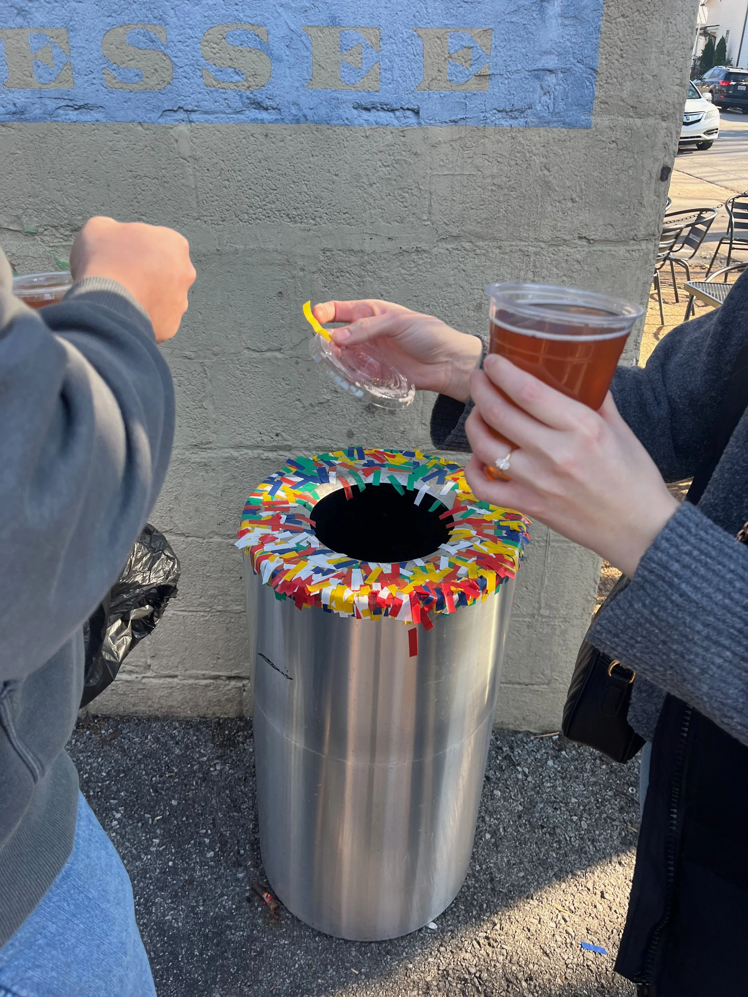 People placing trash into a decorated metal trash can outside, with a wall featuring a partially visible sign reading 'ESSEE' in the background.