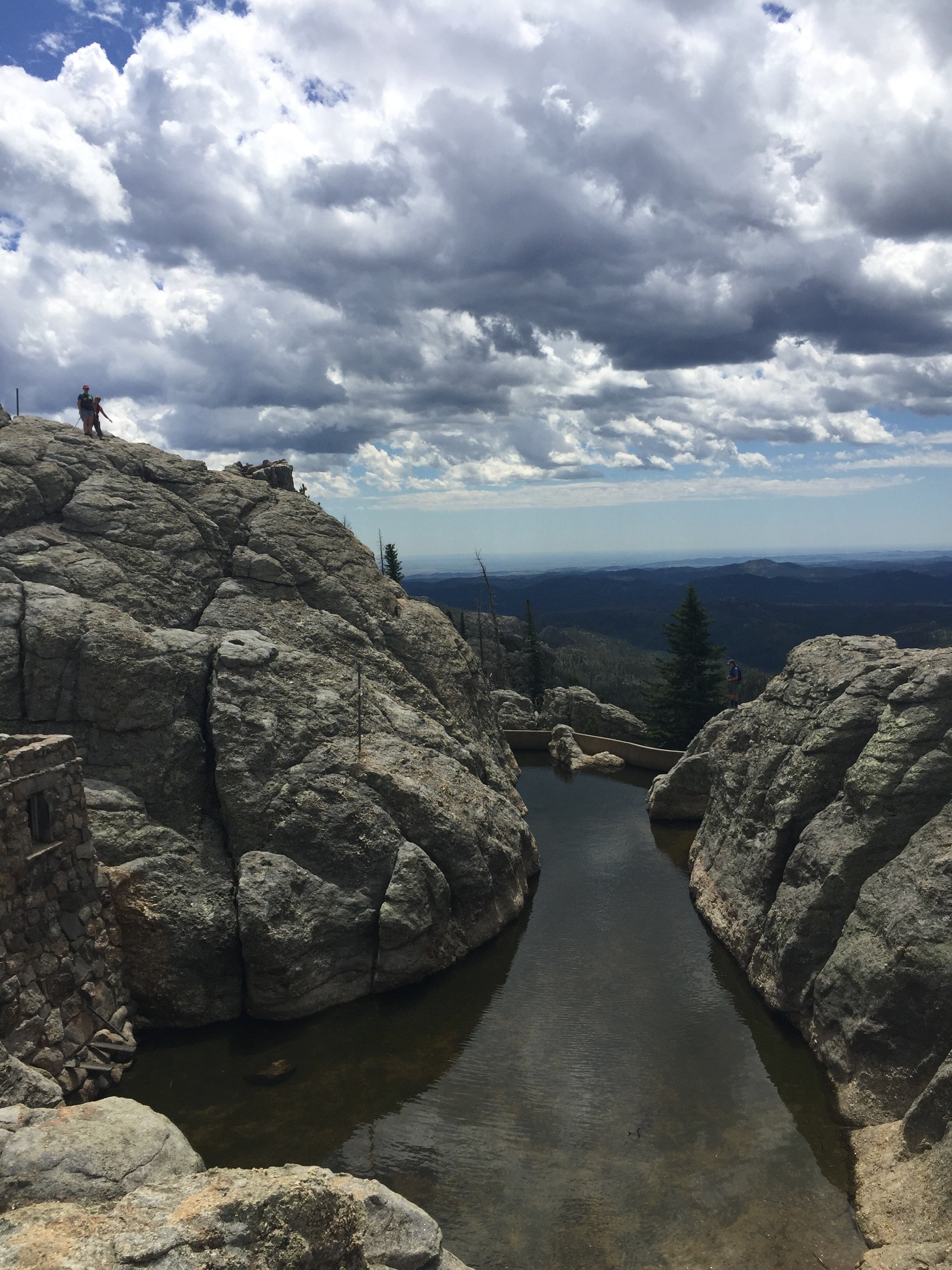 A rocky mountain landscape with a small pool of water and people hiking, under a cloudy sky.