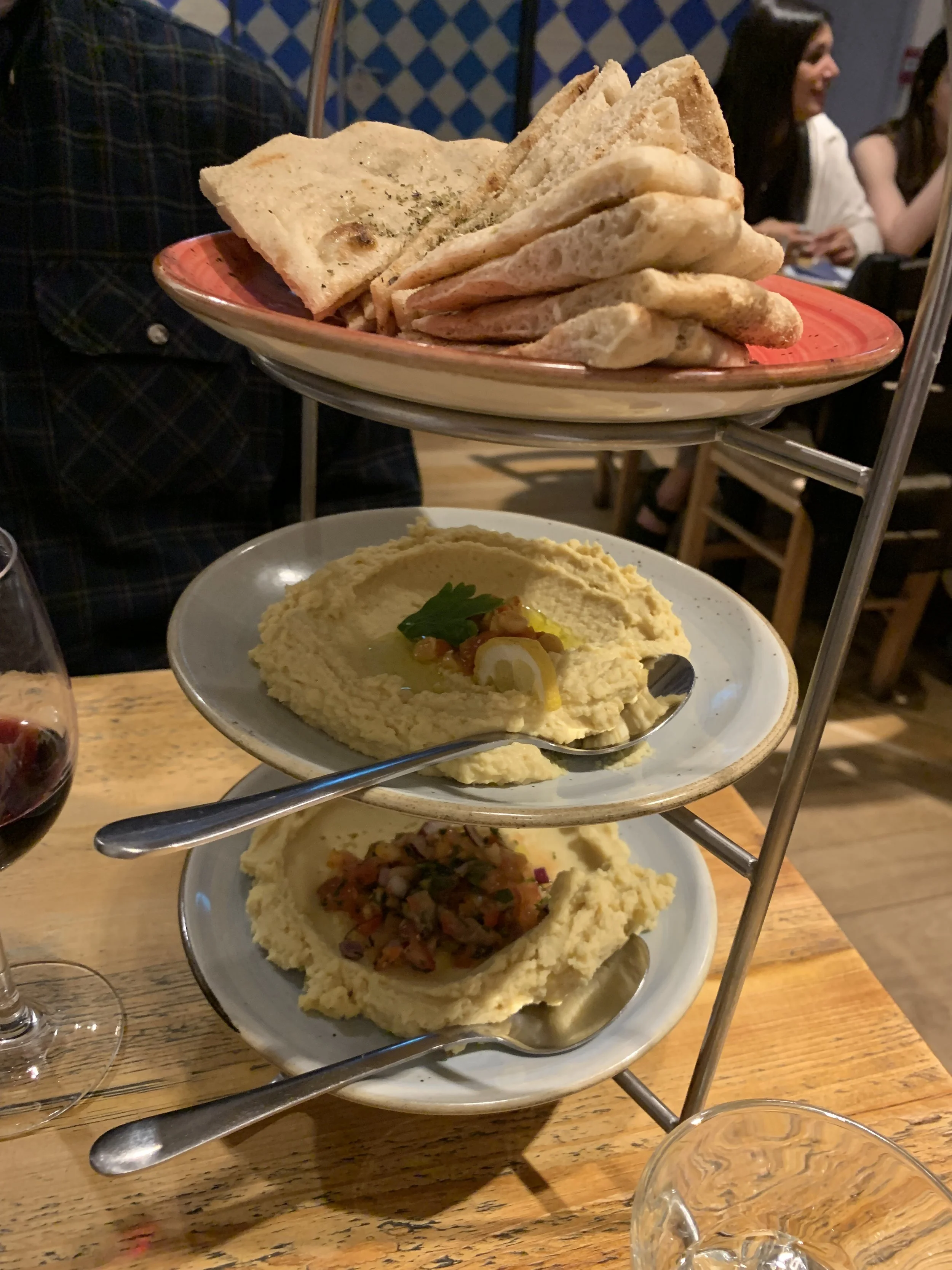 A three-tiered serving stand with sliced bread on the top tier, hummus on the middle tier, and a vegetable dip on the bottom tier, accompanied by glasses of red wine.