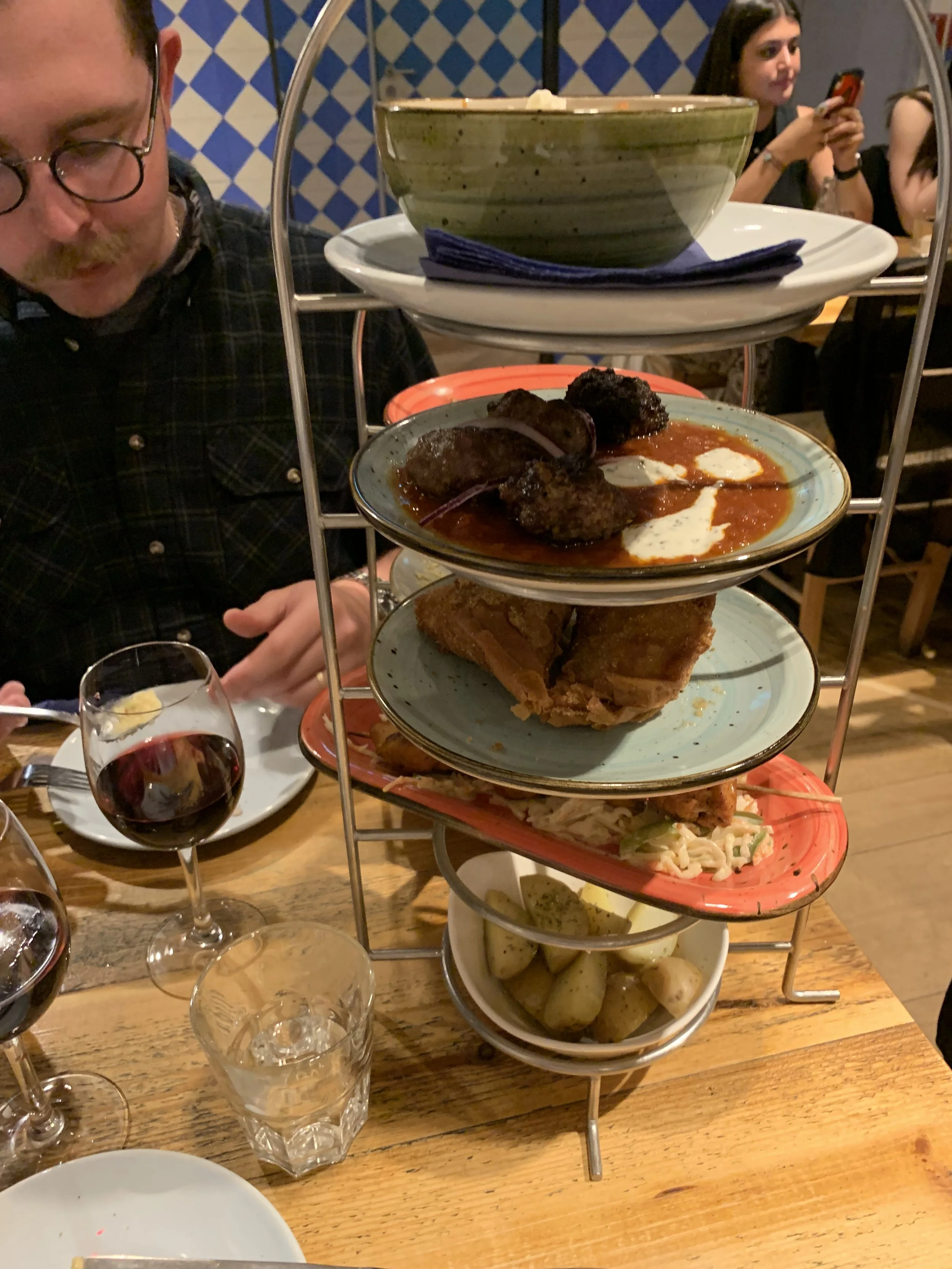 A multi-tiered serving tray filled with various foods such as roasted potatoes, coleslaw, fried dishes, and meatballs, placed on a wooden table at a restaurant with people dining in the background.