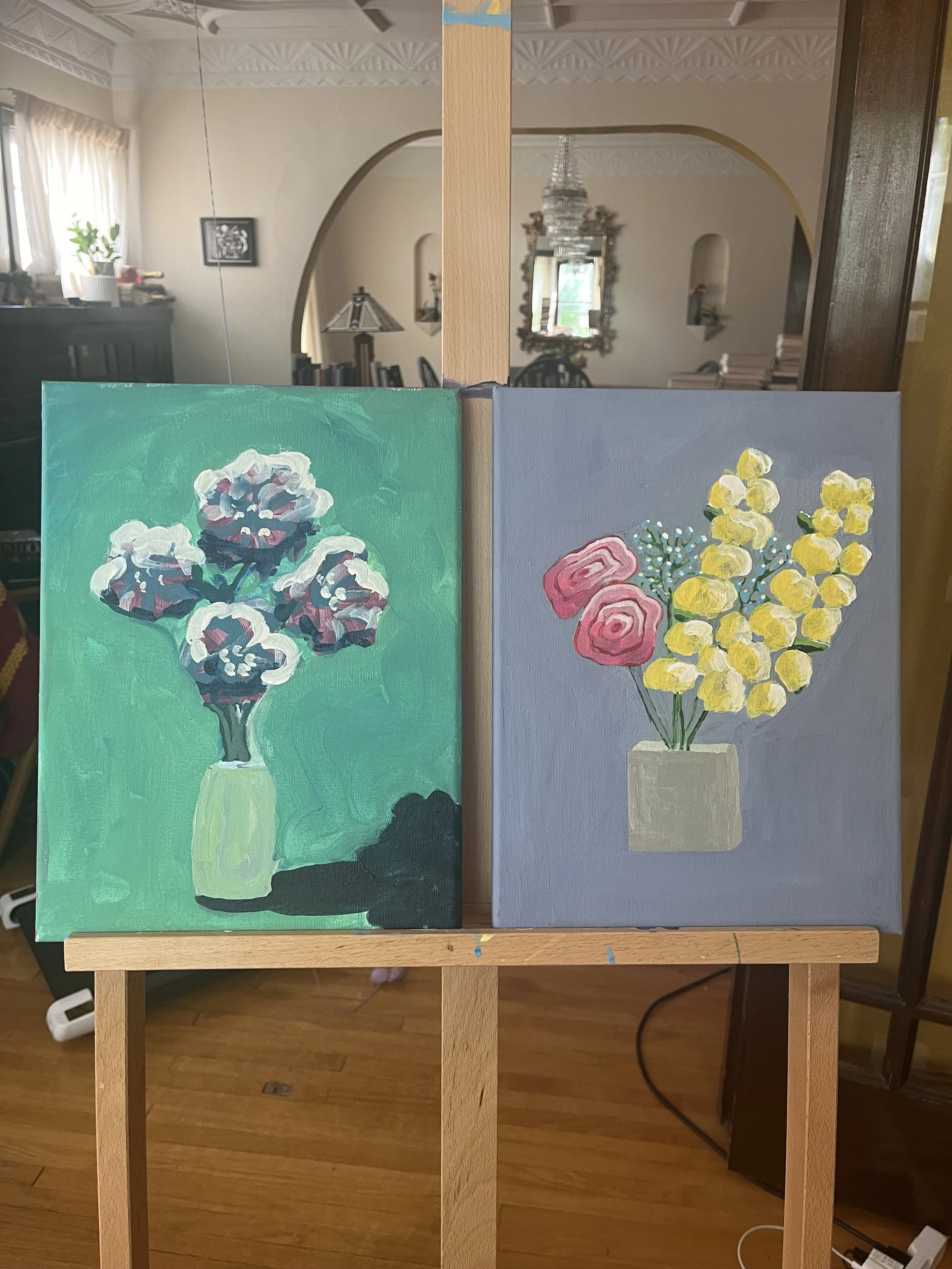Two paintings of flower arrangements on a wooden easel. The left painting features a bouquet of purple and white flowers in a cream-colored vase against a green background. The right painting depicts yellow flowers and pink blooms in a light gray vas