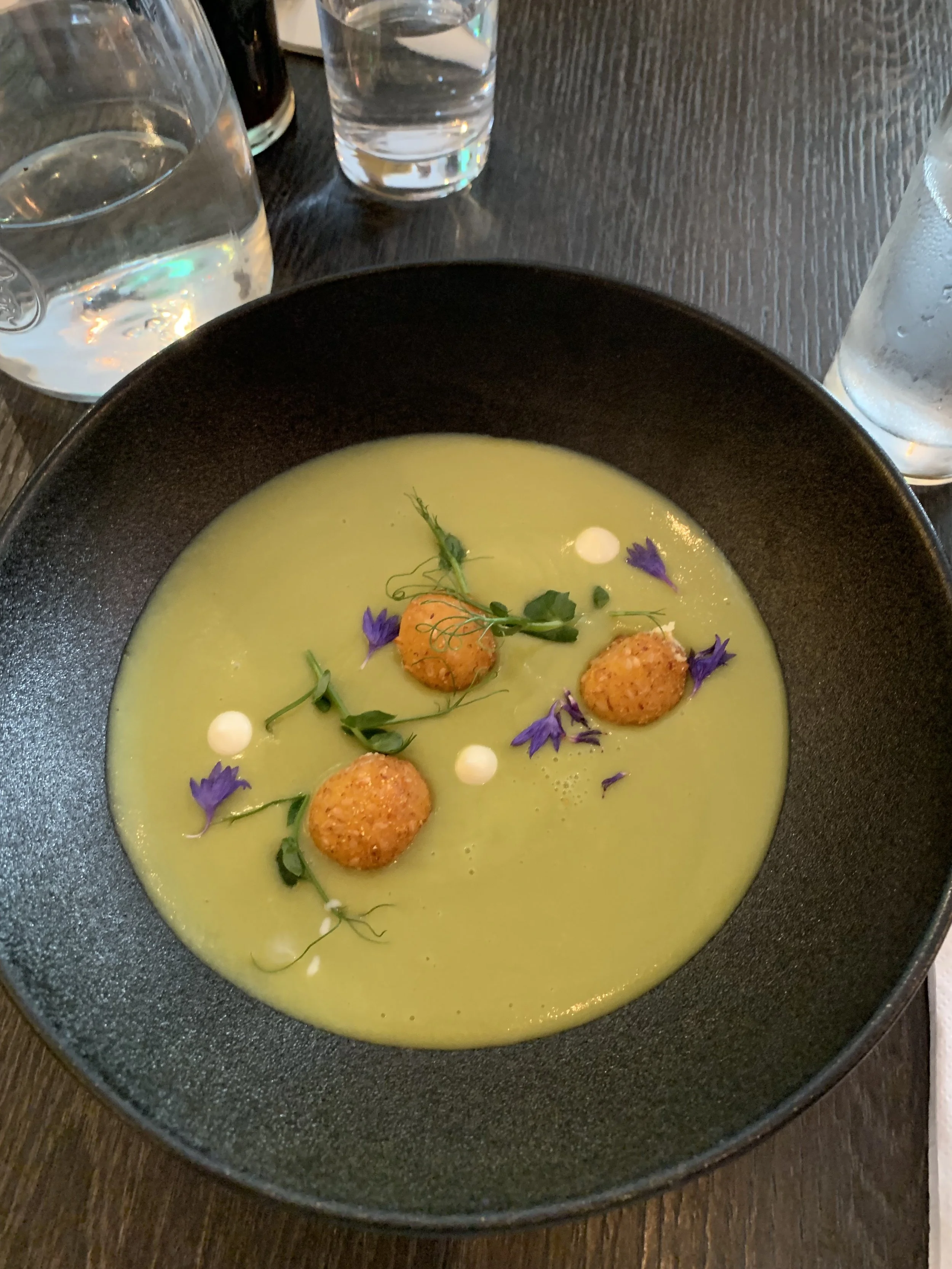 Green soup garnished with small orange croquettes, purple edible flowers, microgreens, and small white dollops in a black bowl on a dark wooden table, with water glasses in the background.