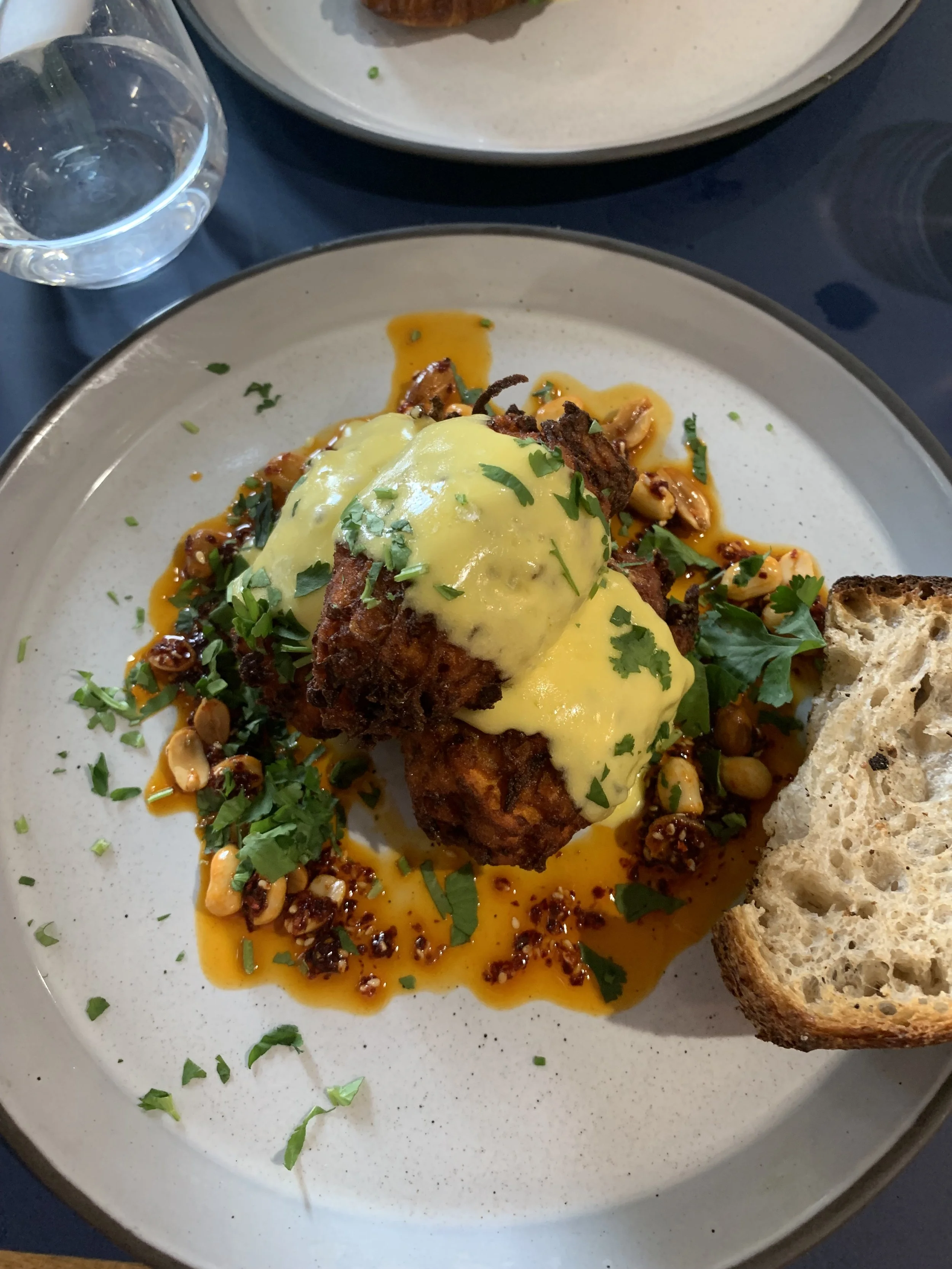 Fried chicken topped with hollandaise sauce, served with a side of toasted bread and garnished with chopped herbs and crushed nuts.