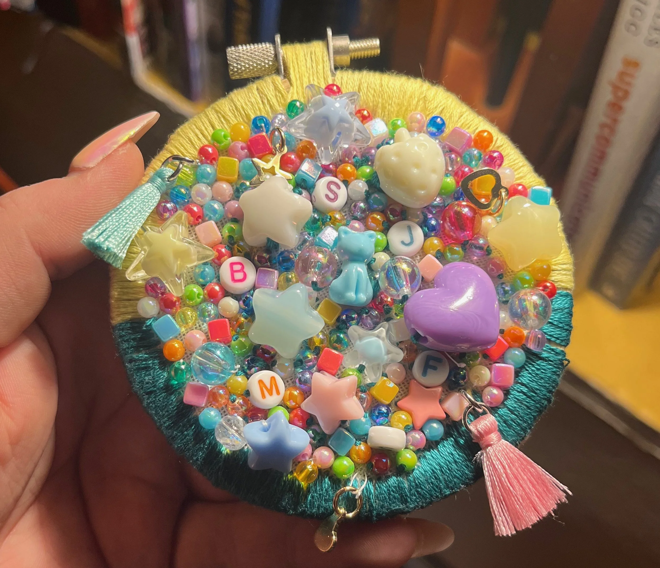 A colorful collection of small jewelry charms and beads, including star, heart, and bear shapes, arranged on a small, round, fabric-covered jewelry holder with tassels at the edges.