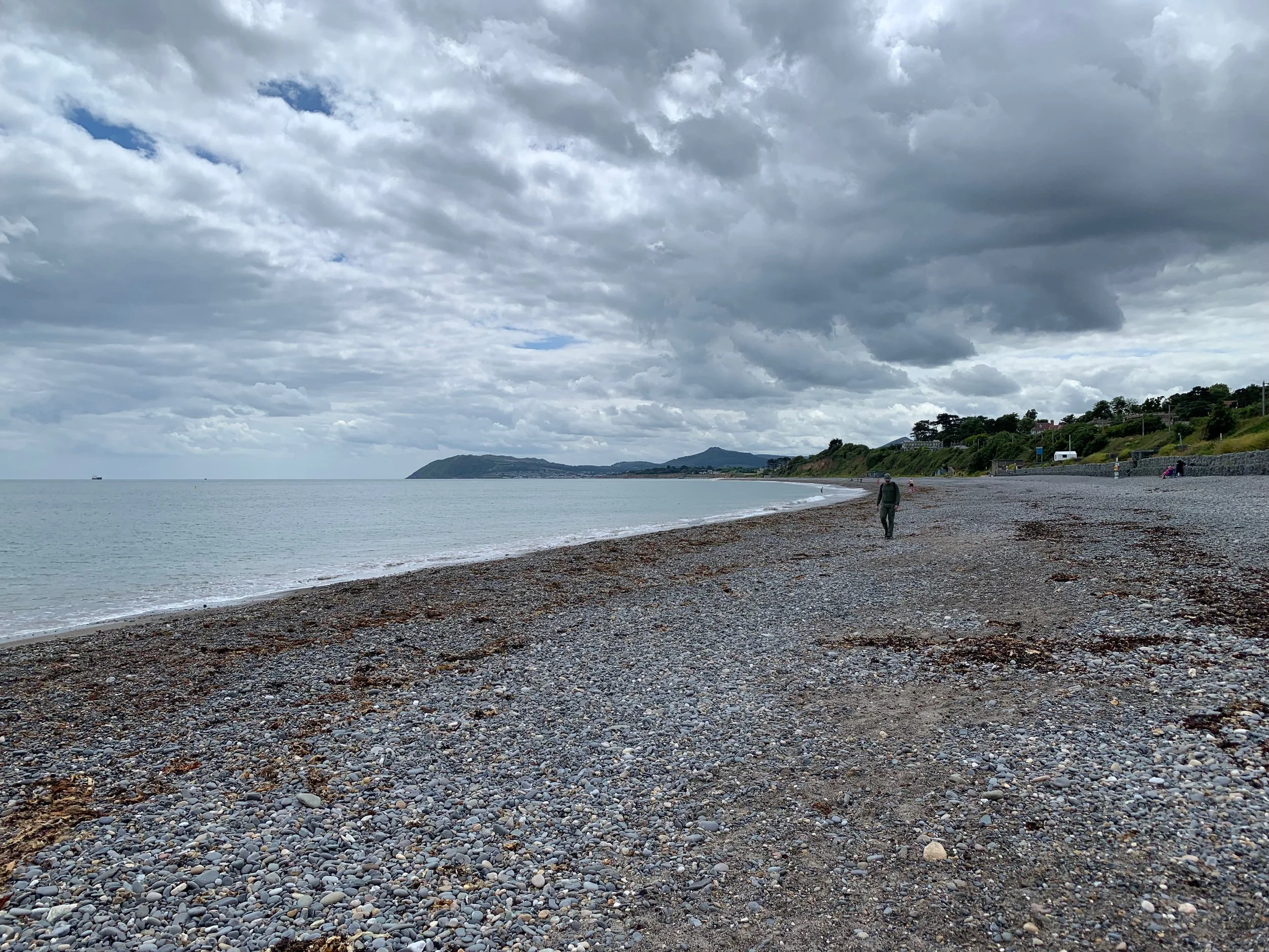 A person walking alone along a pebble-covered beach under a cloudy sky, with hills and trees in the distance.